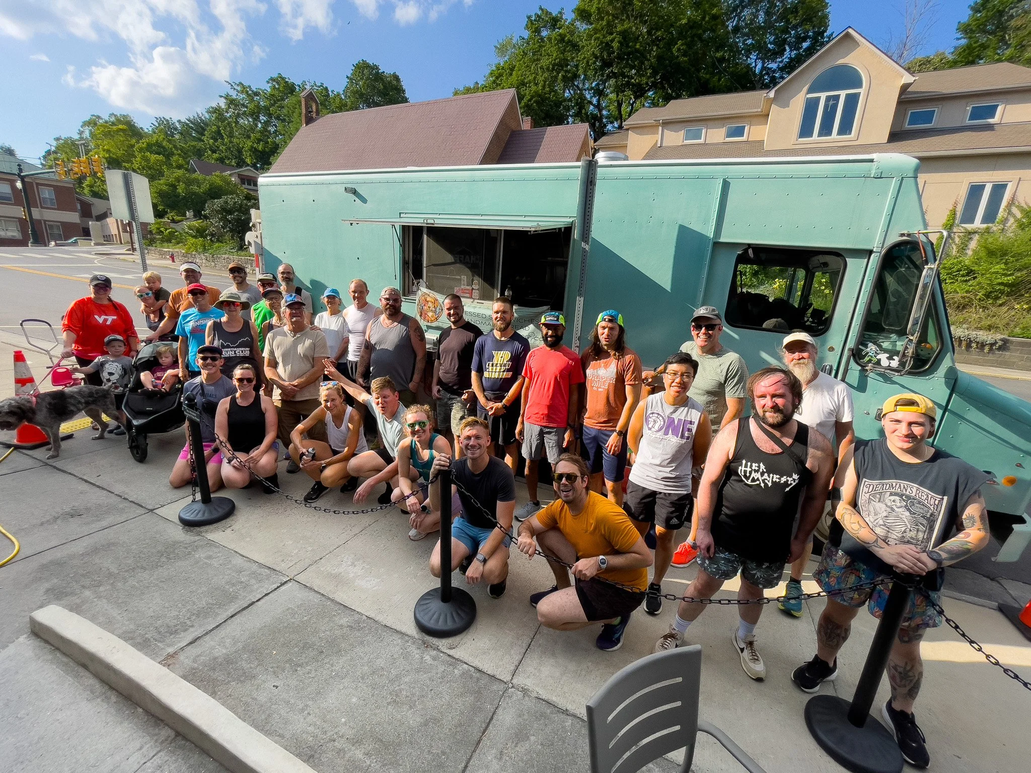 A large group of people standing and kneeling in front of a food truck on a sunny day with green trees and buildings in the background.