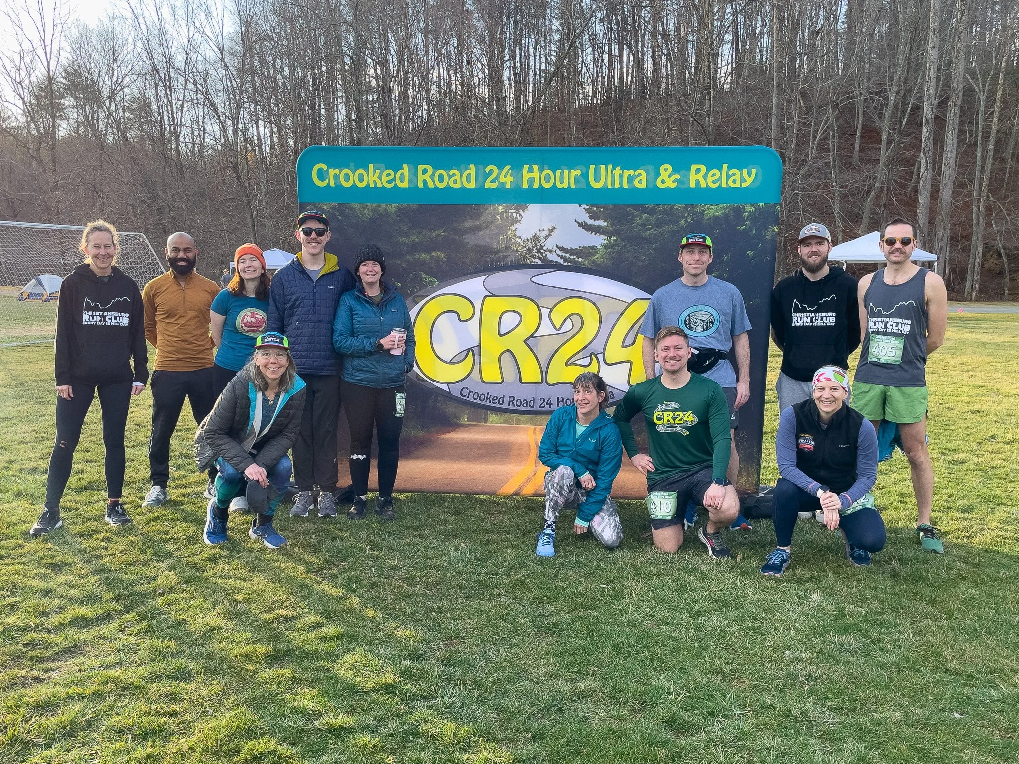 A group of people poses in front of a large sign that reads "Crooked Road 24 Hour Ultra & Relay" and "CR24" with an image of a winding road. The group is outdoors on a grassy field, dressed in athletic clothing, with trees in the background.