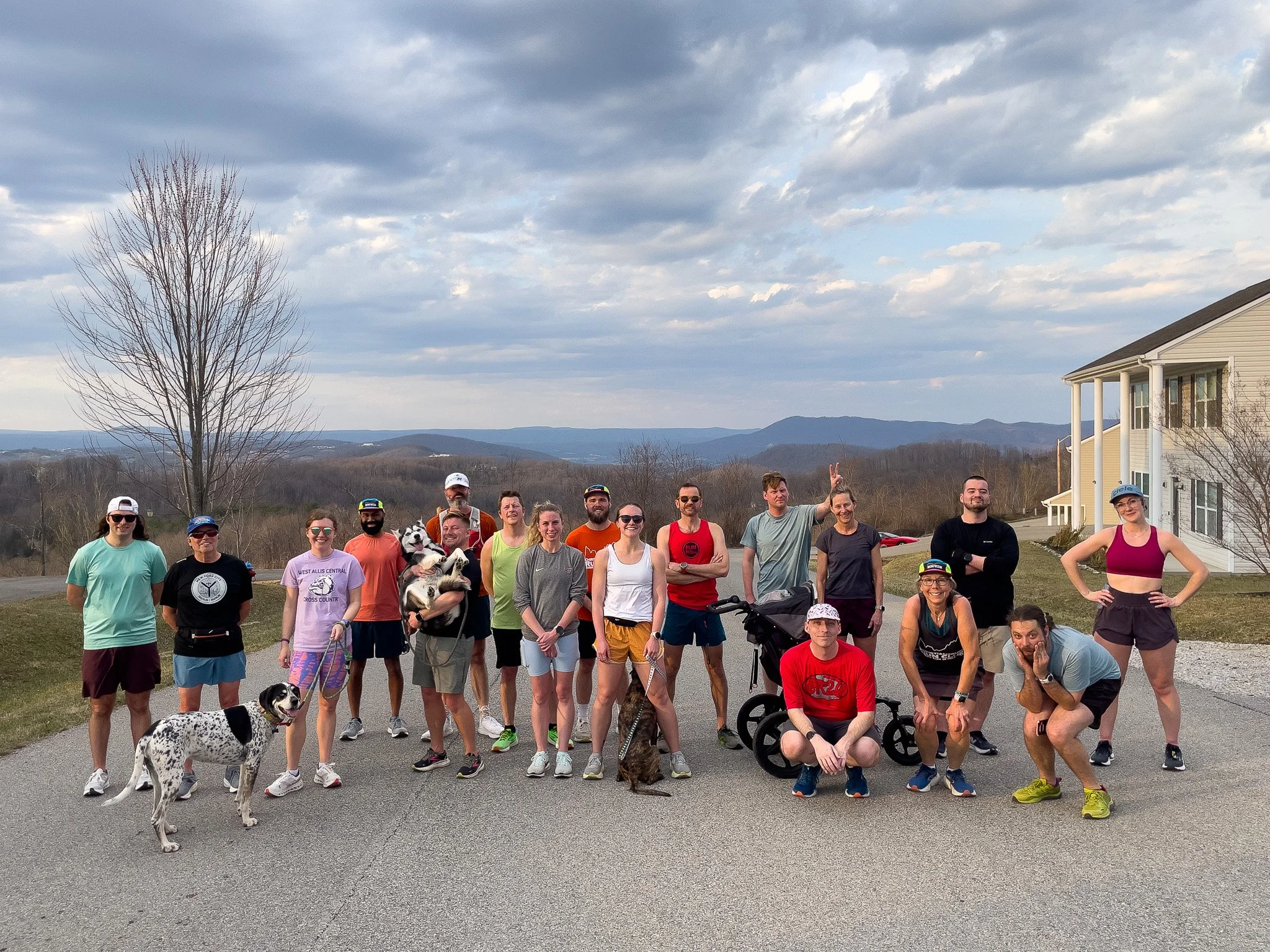 Group of people standing outdoors on a paved surface with scenic mountains and cloudy sky in the background, some with dogs and bicycles.