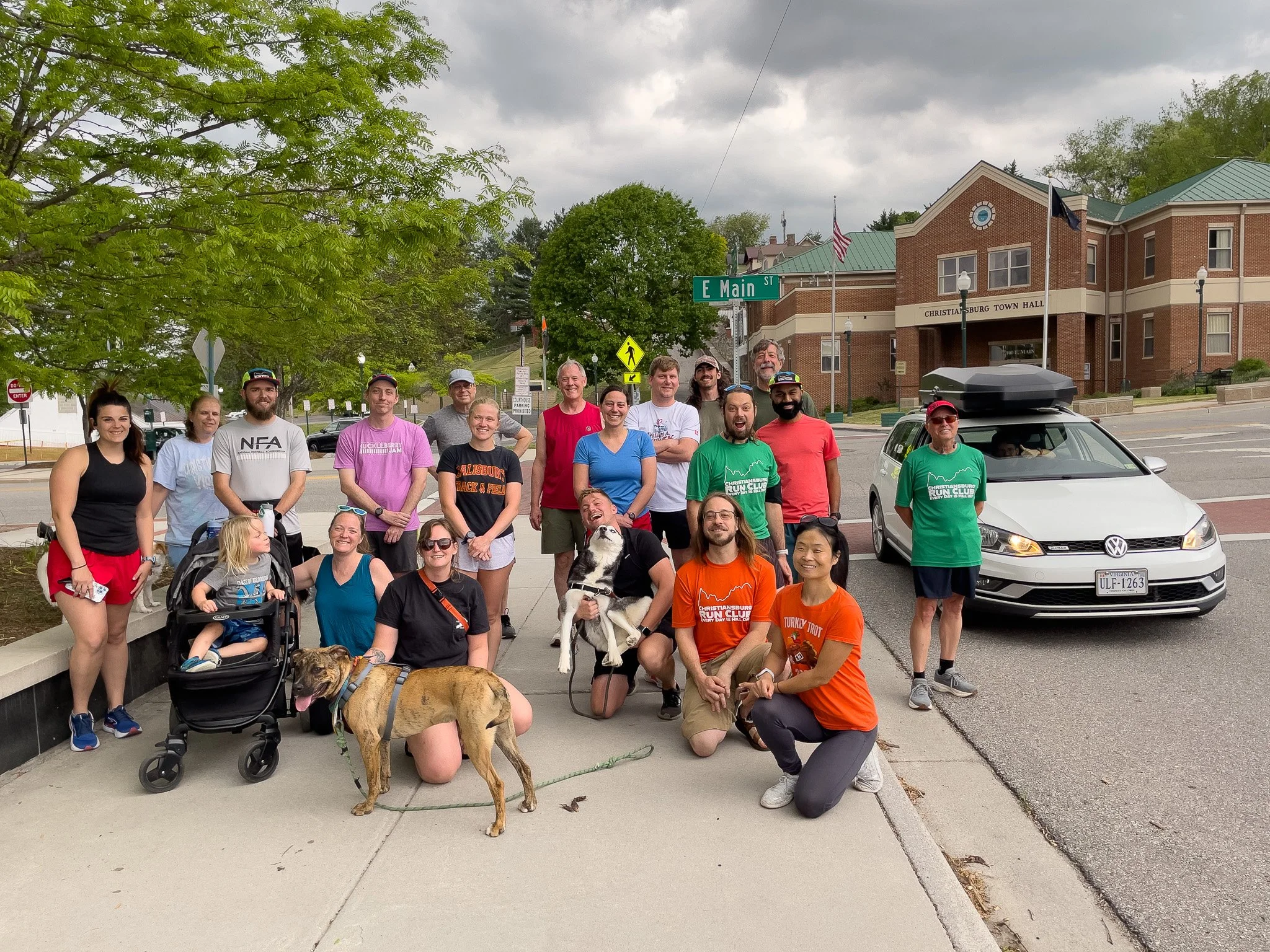 A group of 16 people, including children and dogs, gathered on a sidewalk in front of a brick building with a sign that reads 'Christaensburg Town Hall'. They are smiling and posing for the photo. Some are wearing sports or event T-shirts, and there 