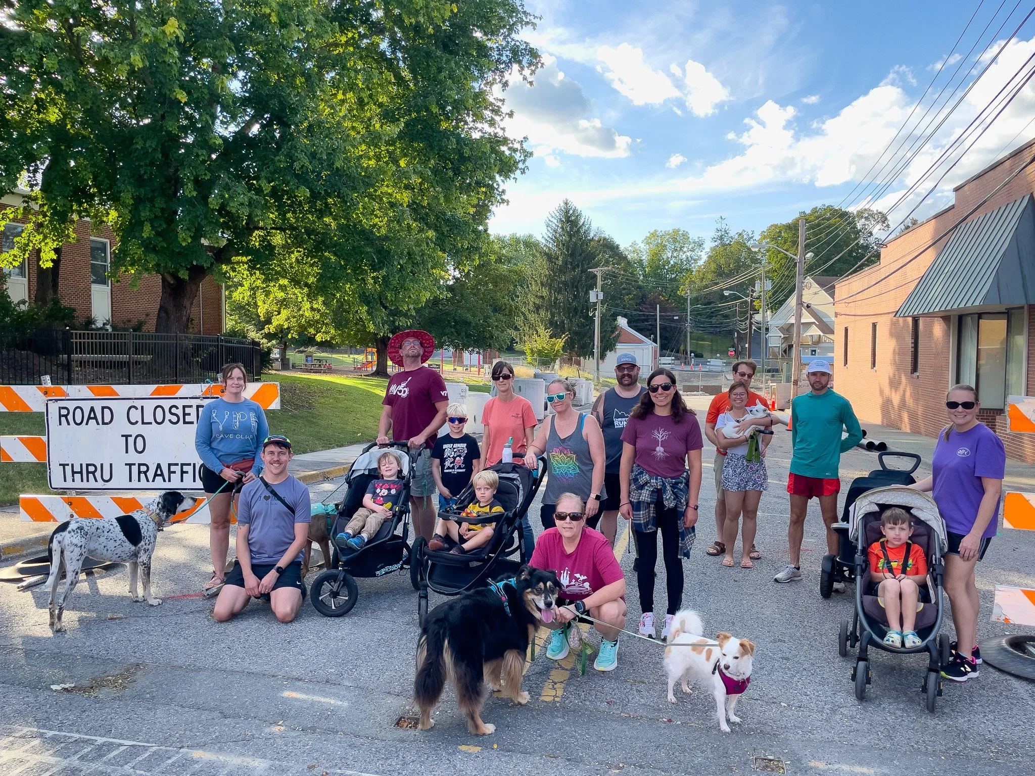 Group of people and dogs gathered in front of a 'Road Closed to Thru Traffic' sign on a street, with trees and buildings in the background, on a sunny day.