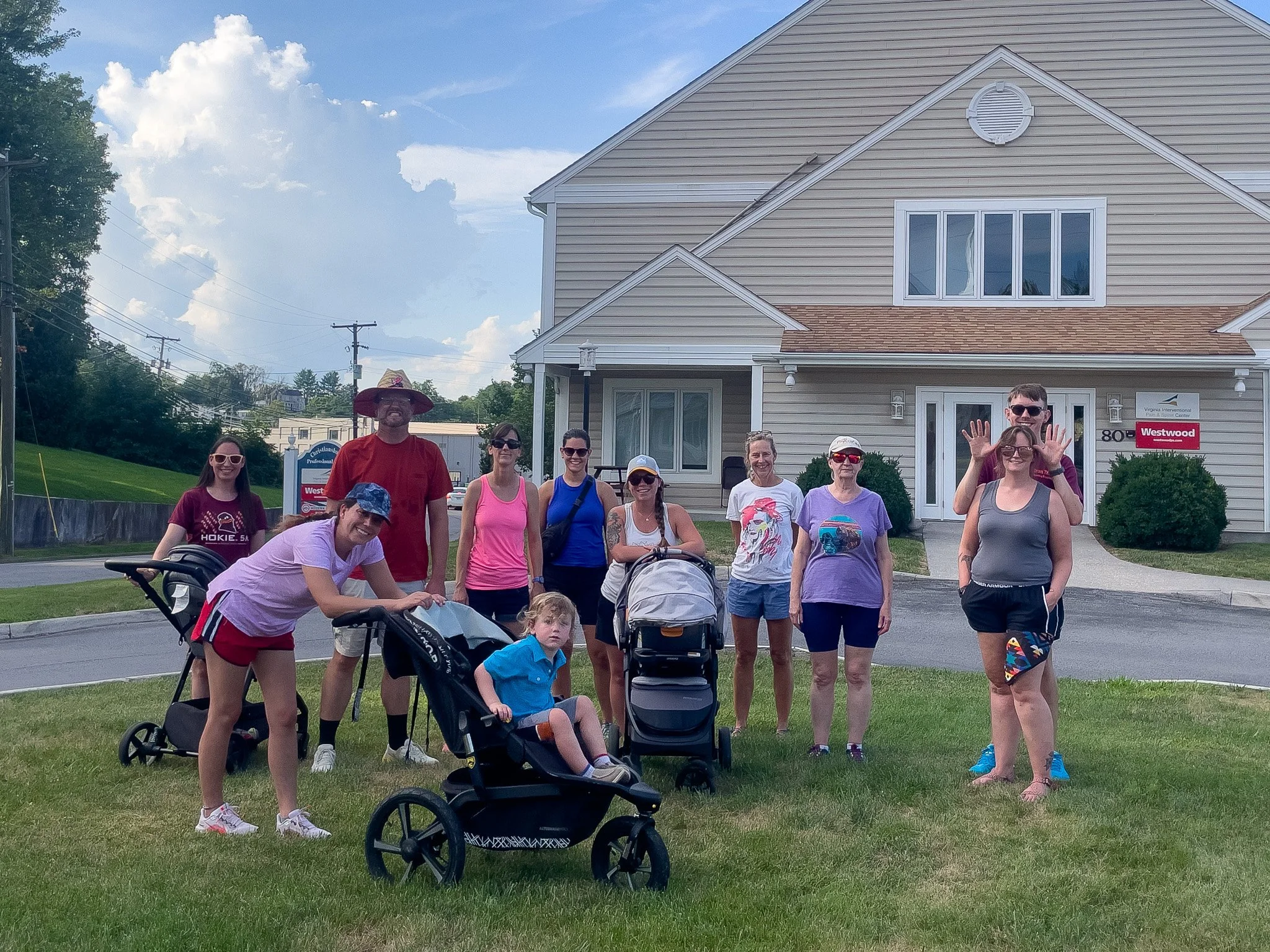 Group of people, including children and adults, standing on a grassy area in front of a beige building with large windows, some in sunglasses and casual sportswear, during daytime with partly cloudy sky.