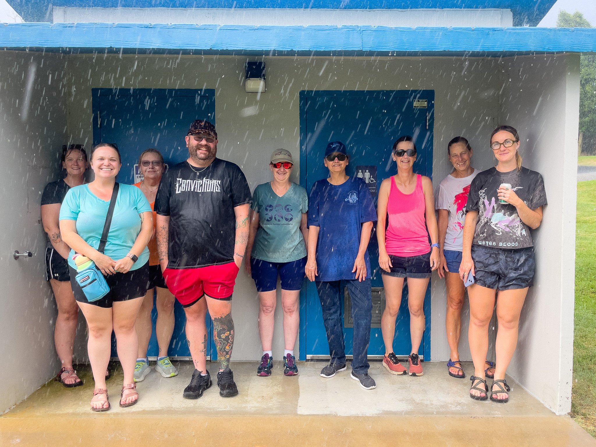 Group of nine people standing under a shelter during rain, smiling and posing for the photo.
