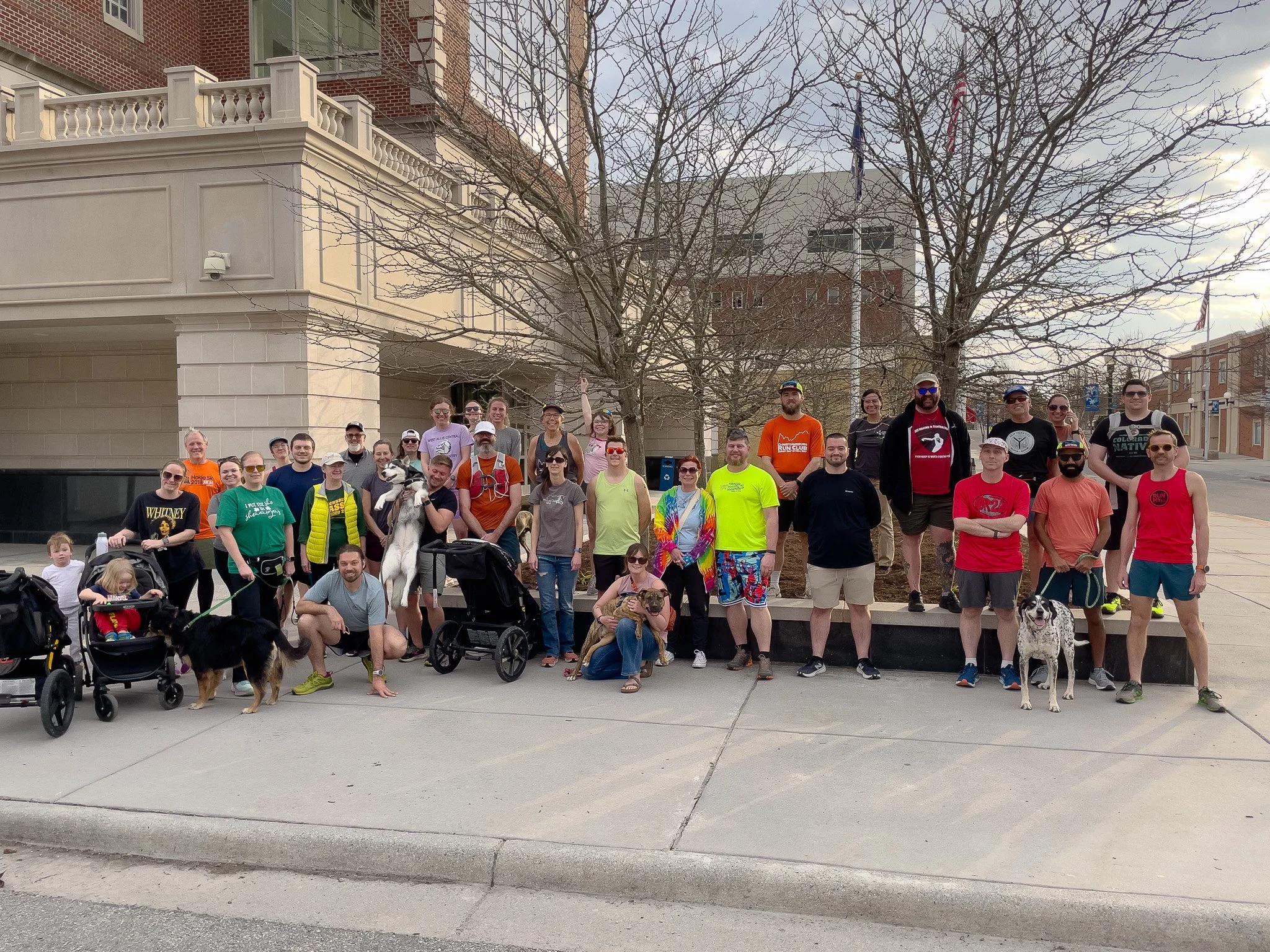 A large group of people, including children and adults, posing outdoors on a city sidewalk. Some are holding leashed dogs and pushing strollers. The background features a building with a tower-like structure and leafless trees.