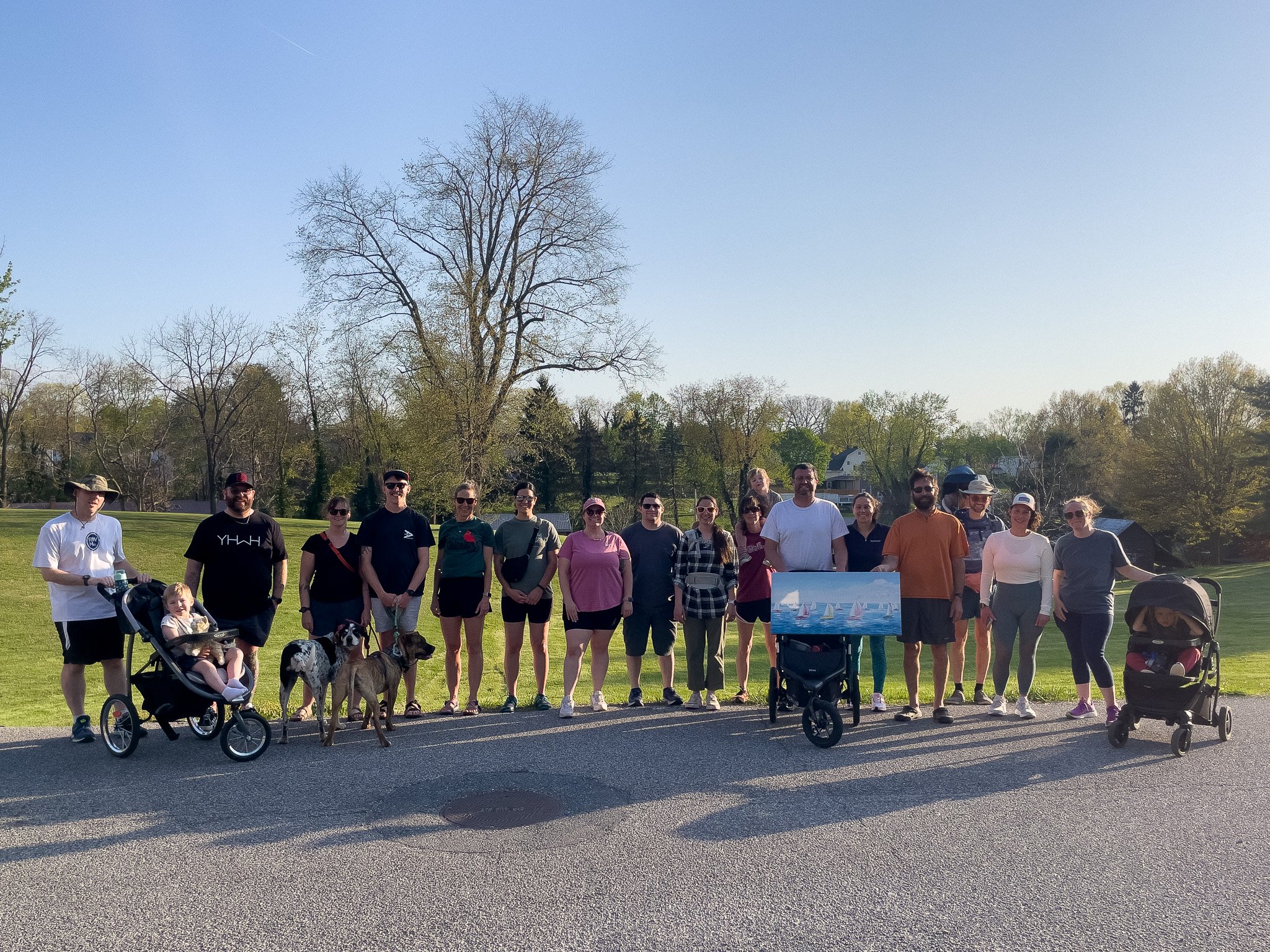 Group of people standing outdoors on a sunny day with trees in the background, some with strollers and dogs.