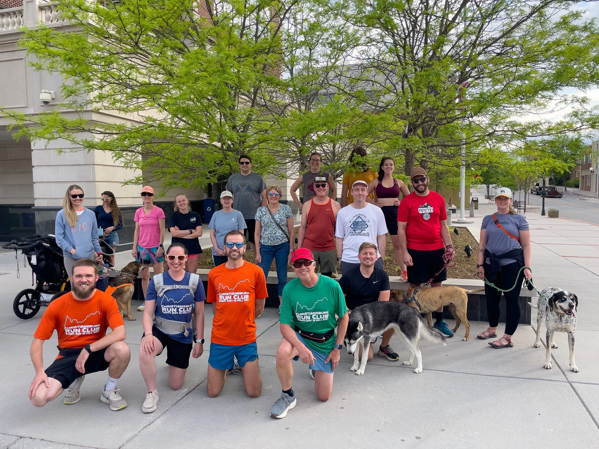 Group of people and dogs gathered outdoors in front of a tree and city building, participating in a running club event.