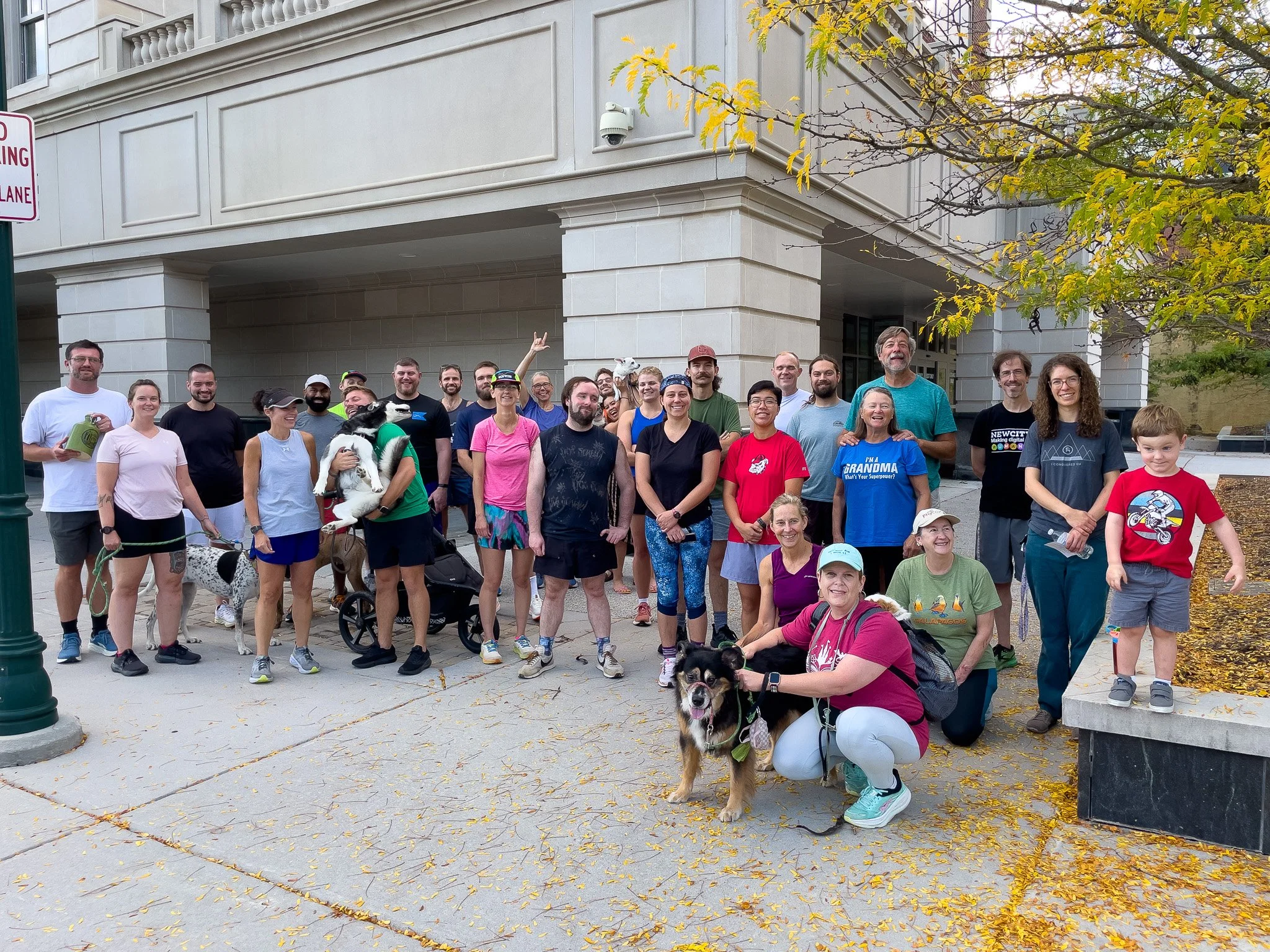 A group of people and dogs posing for a photo outside a building with a tree and fallen leaves in the foreground.