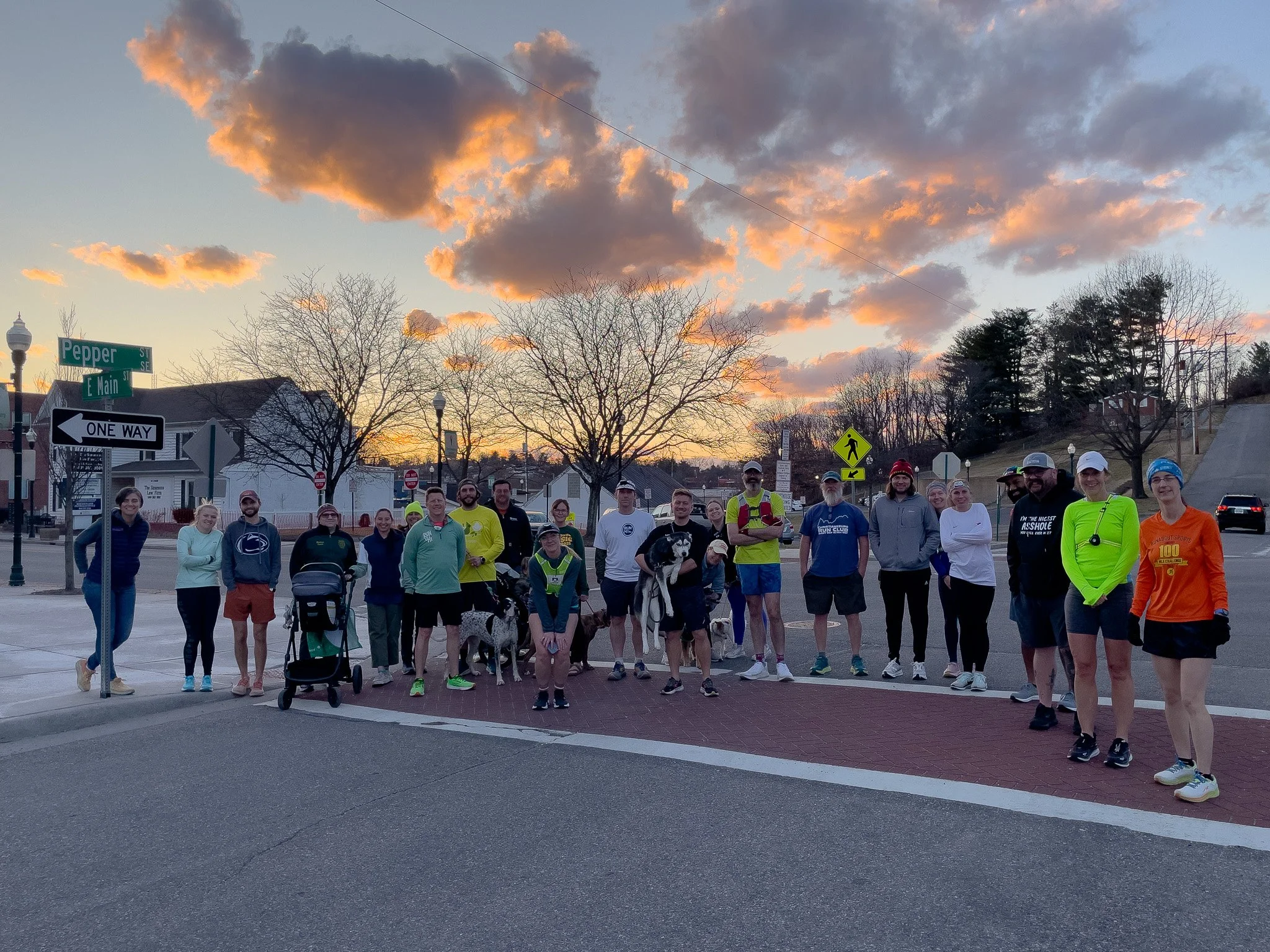 Group of people gathered outdoors at sunset, standing near street signs at the corner of Pepper Street and E Main Street, some with dogs, wearing running and athletic clothing.