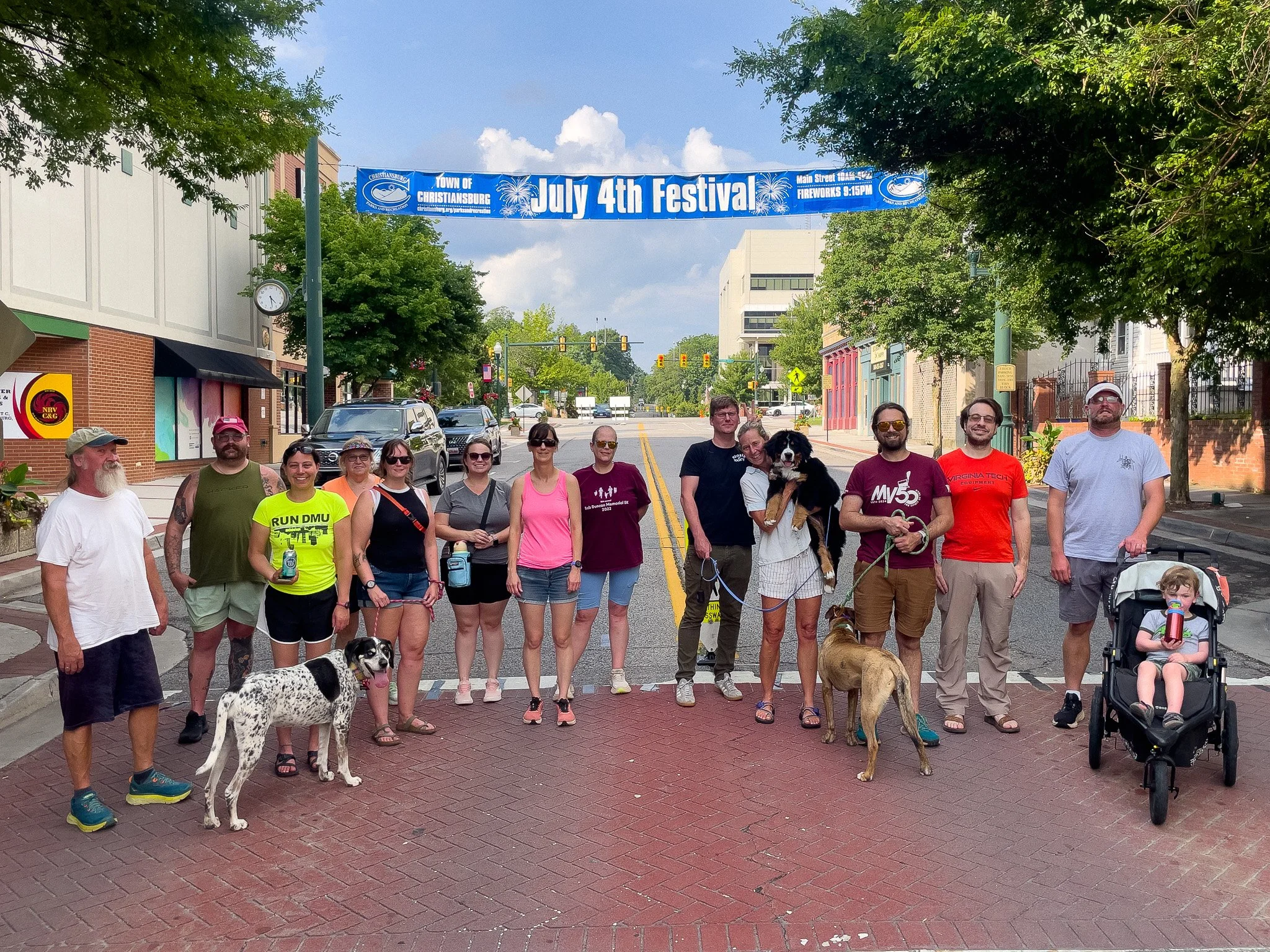 Group of people and dogs standing on a brick street under a banner that reads 'July 4th Festival' in a downtown area.