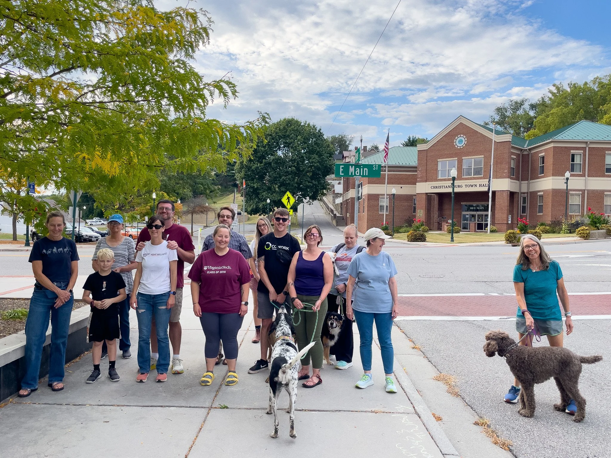 A group of people and dogs standing on a sidewalk at an intersection in front of the Christiansburg Town Hall in Virginia, with a green tree and blue sky in the background.