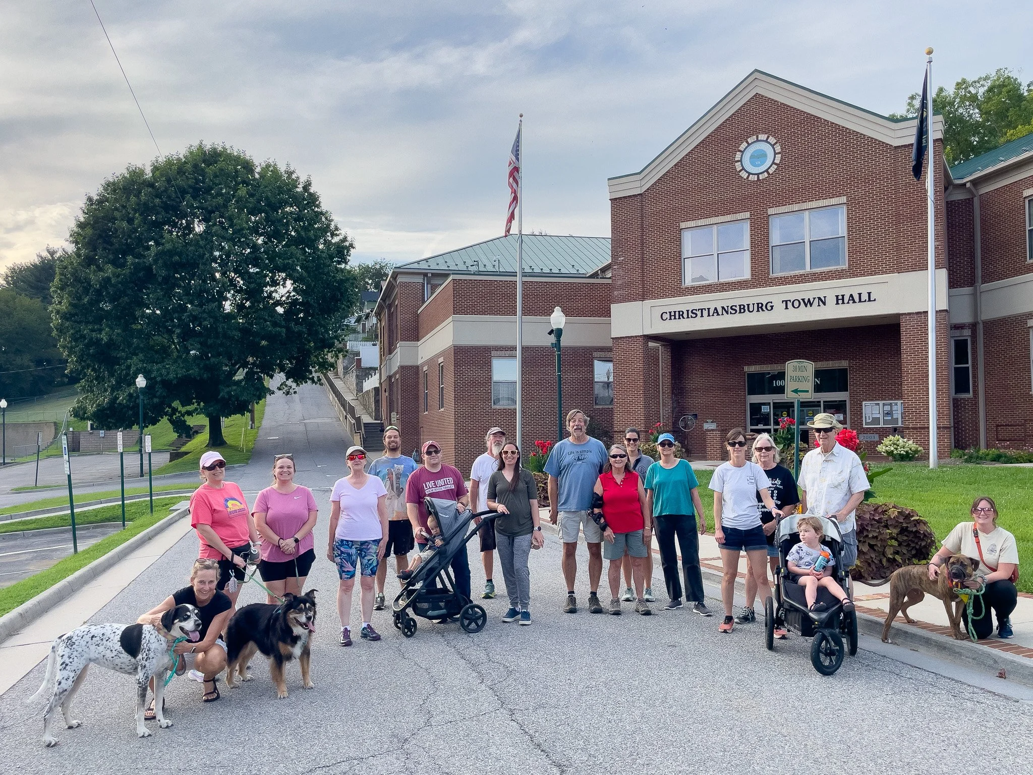 Group of people with dogs standing in front of Christiansburg Town Hall with a large tree and a hill in the background.