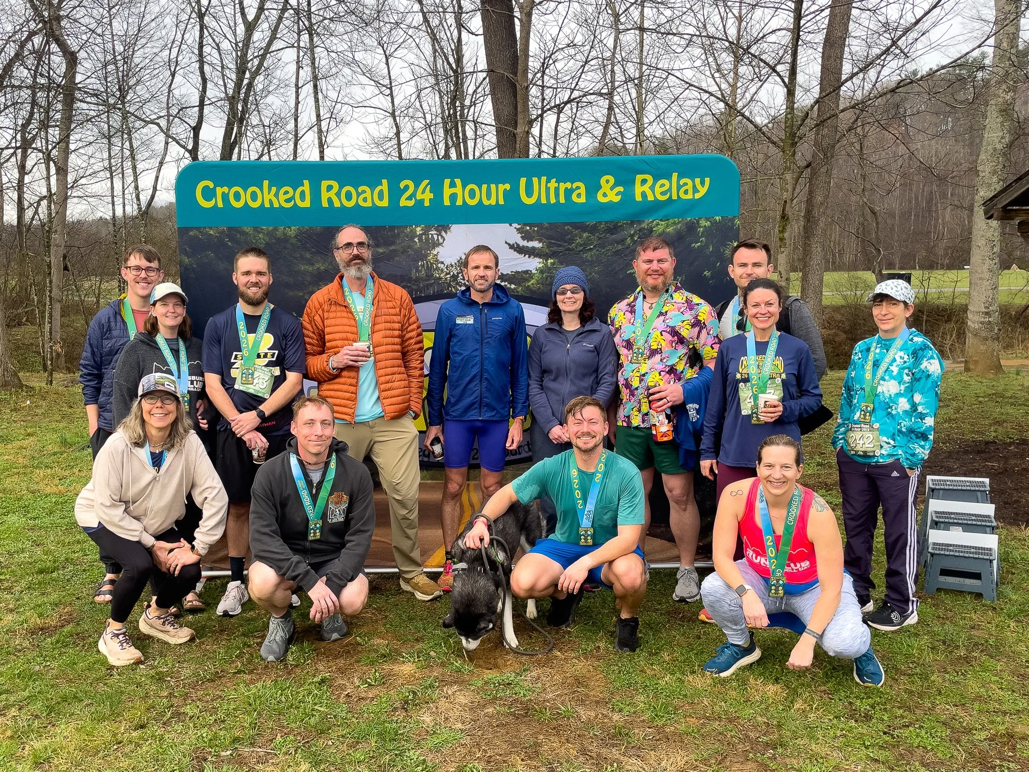 Group of people posing outdoors in front of a sign that reads "Crooked Road 24 Hour Ultra & Relay." They are wearing medals and athletic gear, with some holding drinks, and a dog is in front of the group.