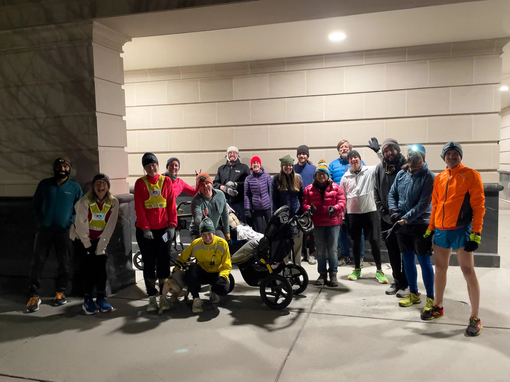 Group of people dressed in running and athletic gear, standing outside at night, some smiling and waving, with a stroller and a dog among them.