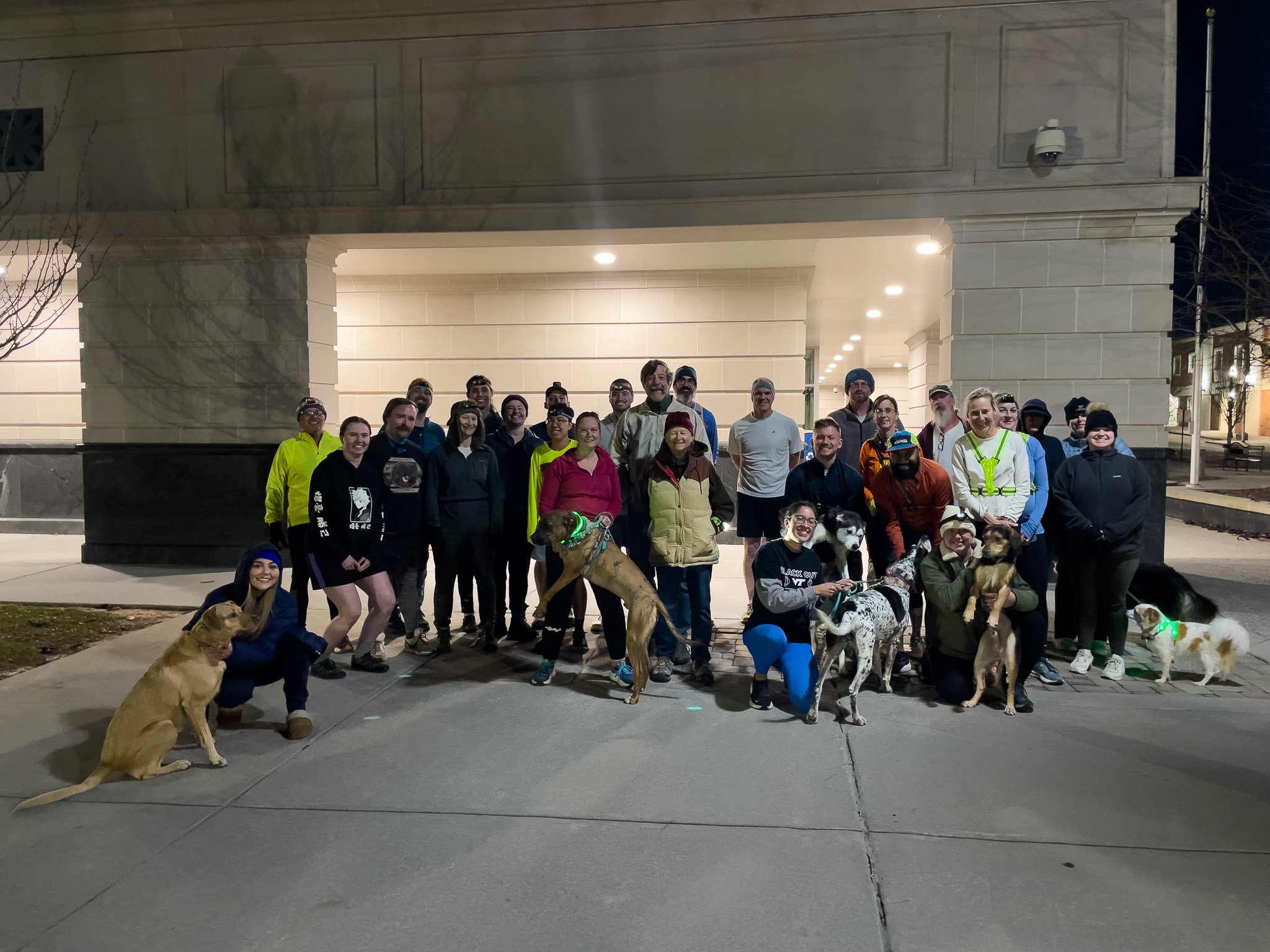 Group of people with dogs gathered outside at night in front of a building, smiling for the camera.