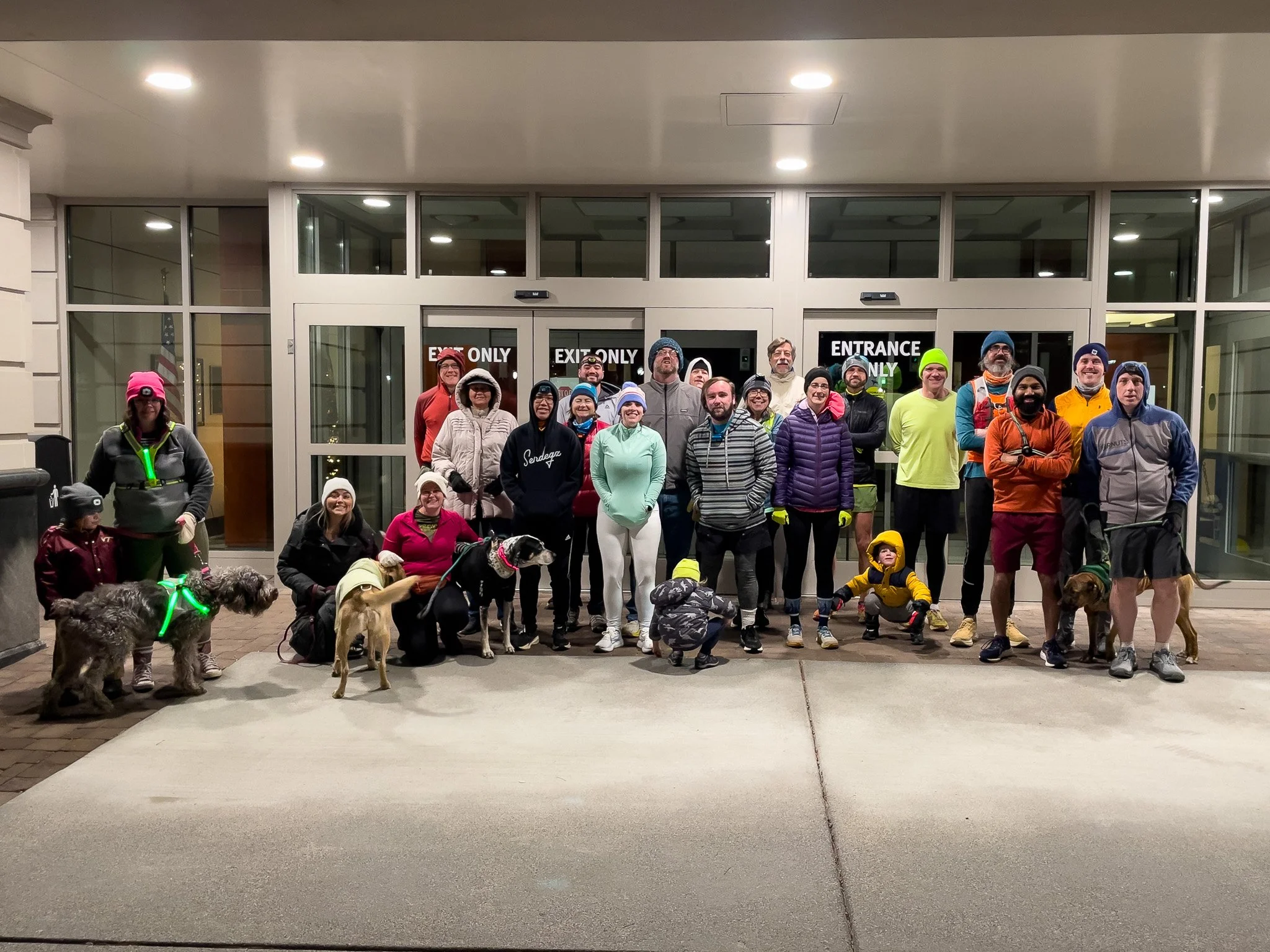 Group of people and dogs standing in front of a building entrance at night, dressed in athletic wear suitable for cold weather.