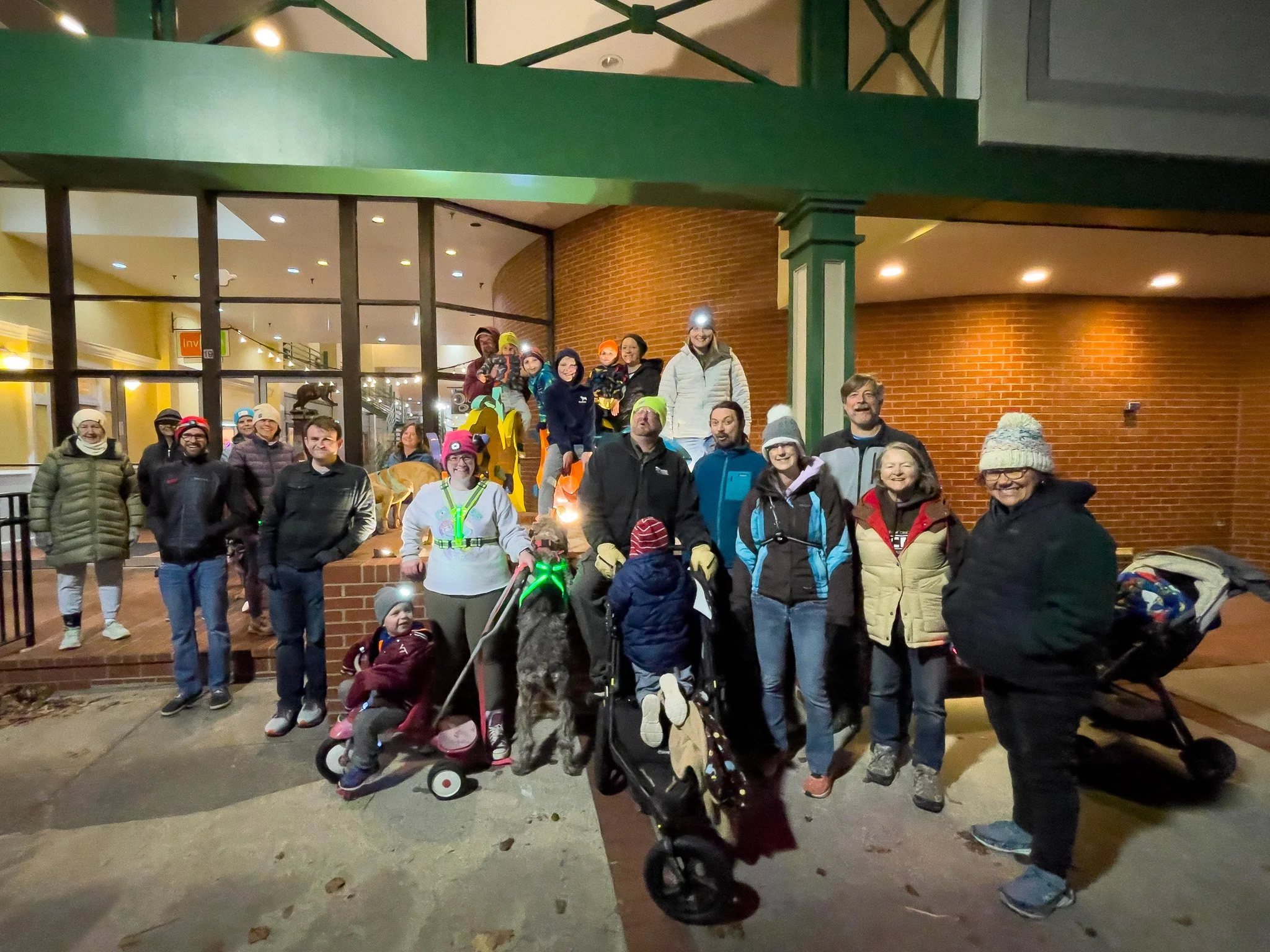 A group of people, including children and adults, gathered outdoors at night, posing for a photo in front of a building with brick walls and large windows. Some are dressed warmly in jackets, hats, and gloves, and there is a dog on a leash with a gre