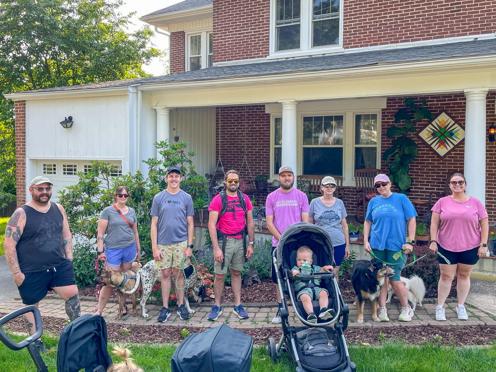 A group of people and dogs posing for a photo in front of a brick house with a porch, trees, and plants.