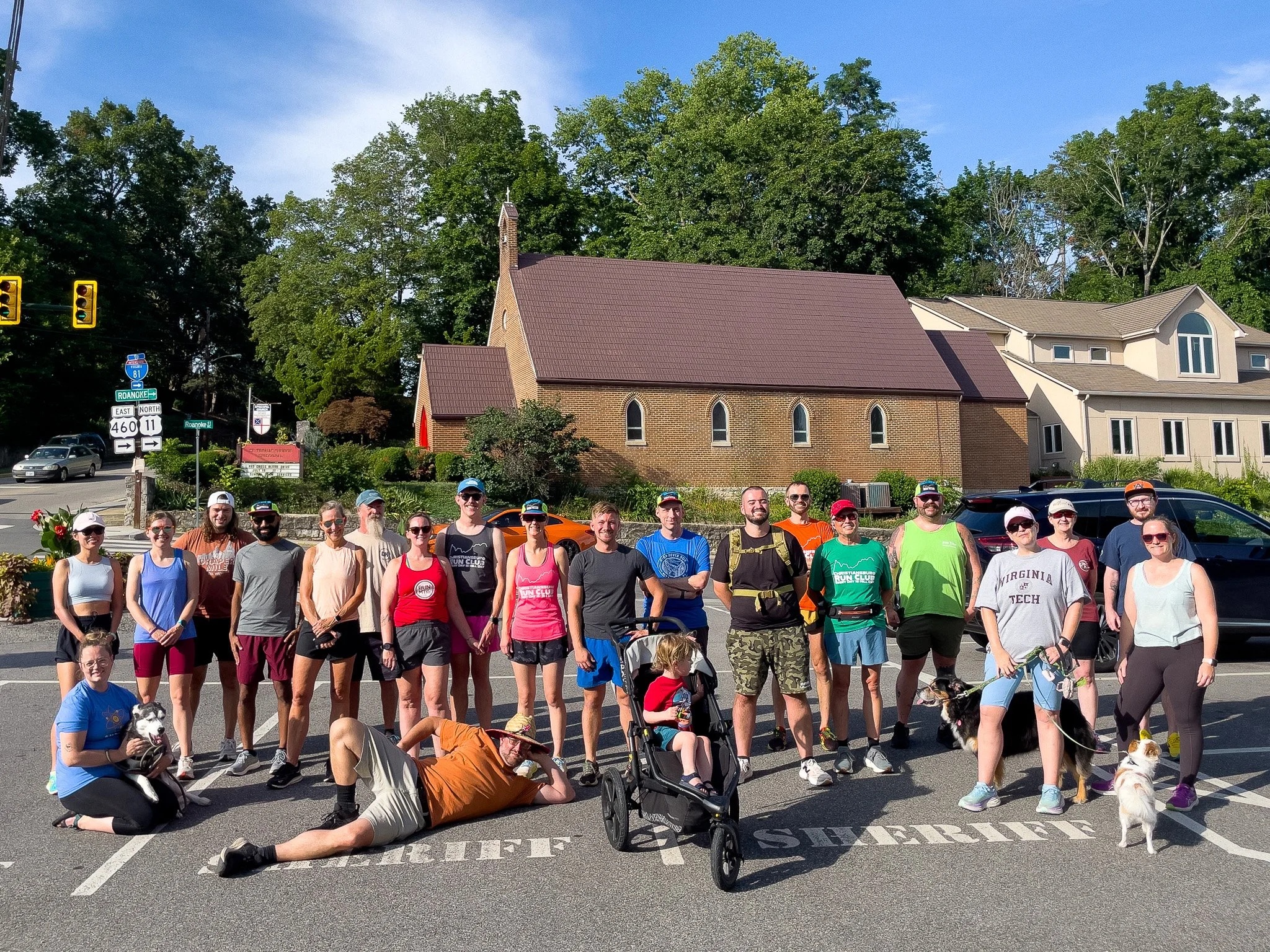 Group of people with their dogs and a stroller standing and sitting on a parking lot marked with 'STAFF' and 'RESERVE' on a sunny day.