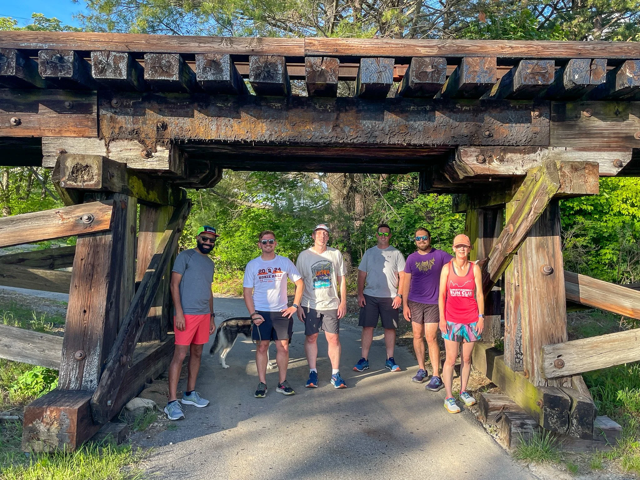 Group of six people standing under a rustic wooden bridge on a trail, surrounded by green trees.