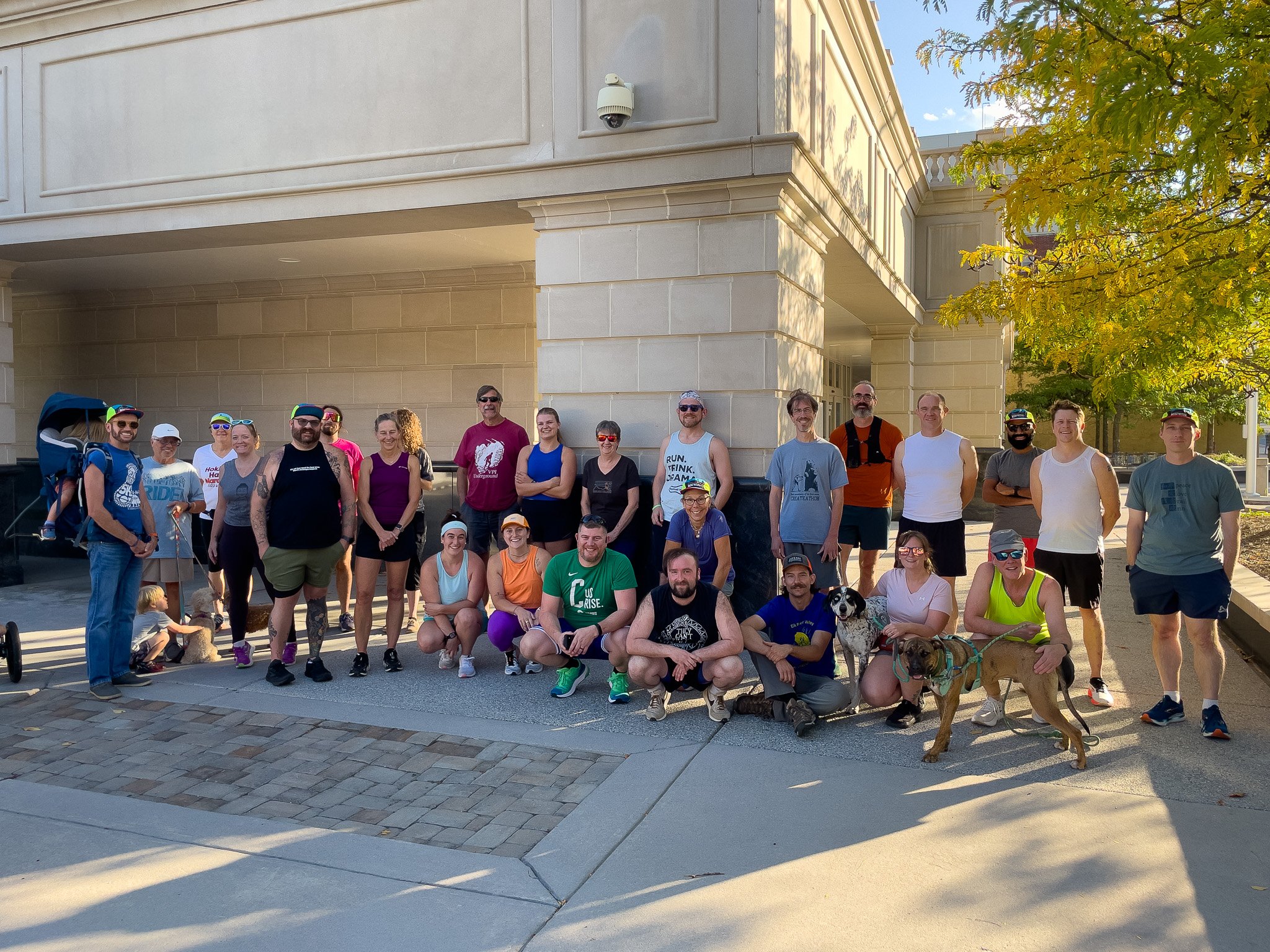 Group of people, dressed in athletic clothing, standing and kneeling outdoors in front of a tan building, with some dogs and a stroller, during daytime.
