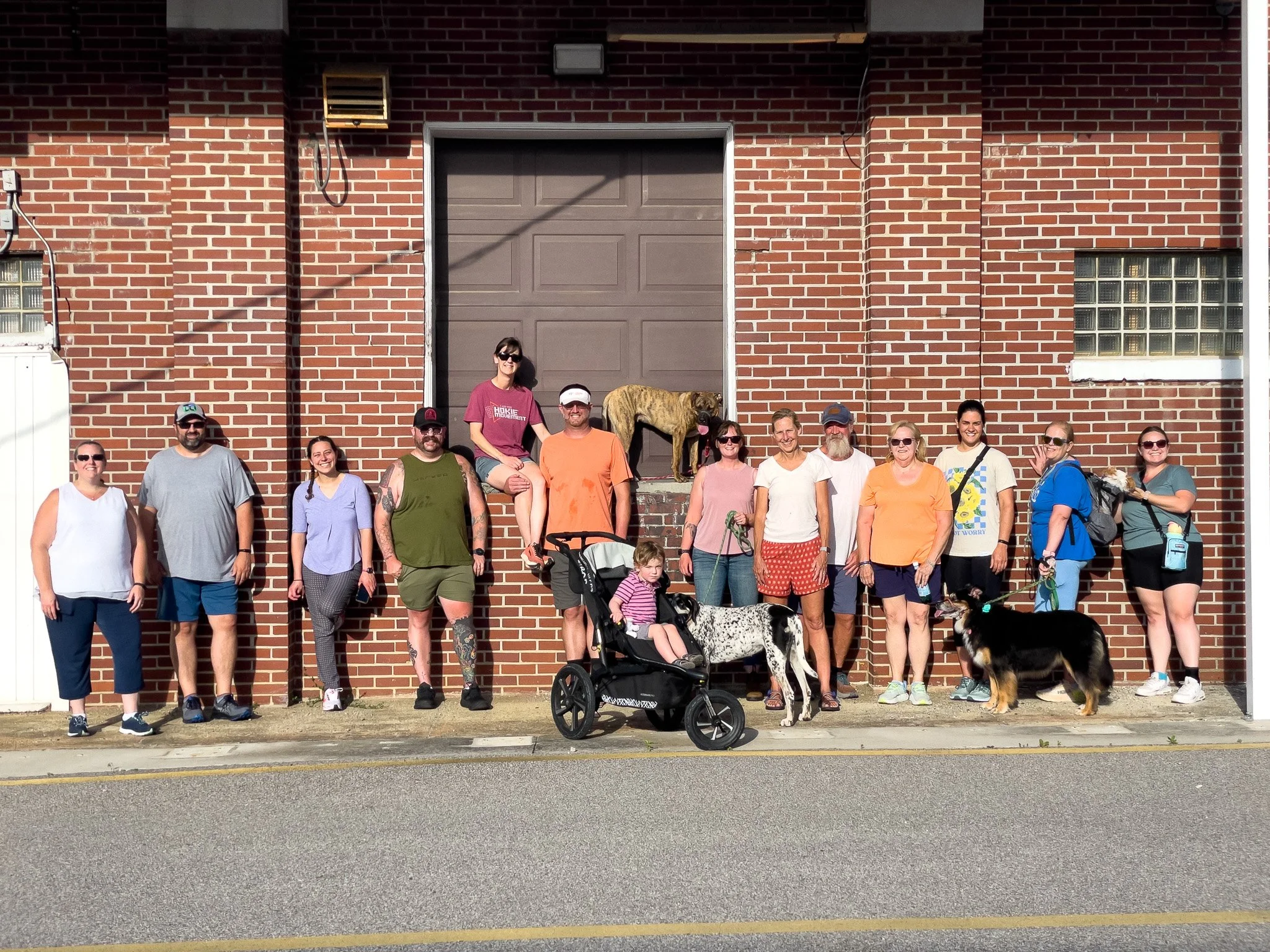 Group of people and dogs standing and sitting in front of a brick building with a raised dog statue.