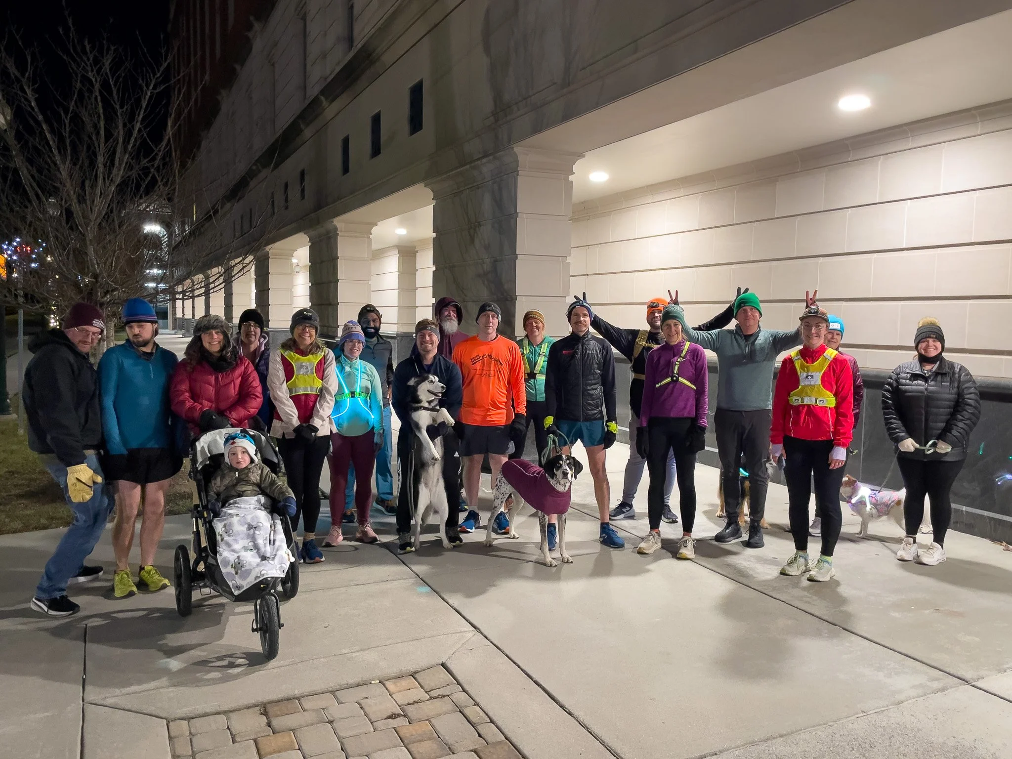 Group of people, including adults and children, standing outdoors at night, dressed in athletic clothing and reflective safety gear, some making peace signs, with dogs and a stroller, in front of a large building with front porch lights.