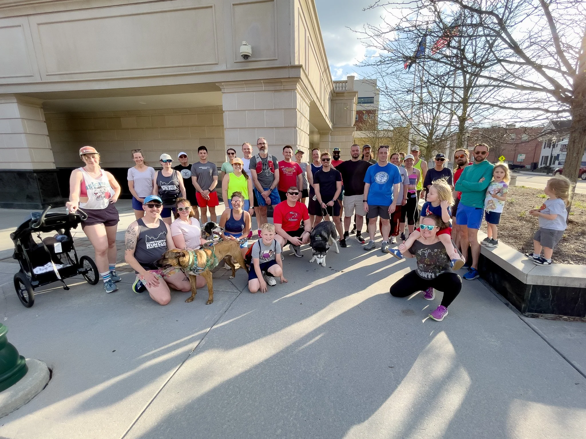 Group of people of various ages and genders, some with dogs and children, gathered outdoors on a sunny day in front of a building with leafless trees and a clear sky.