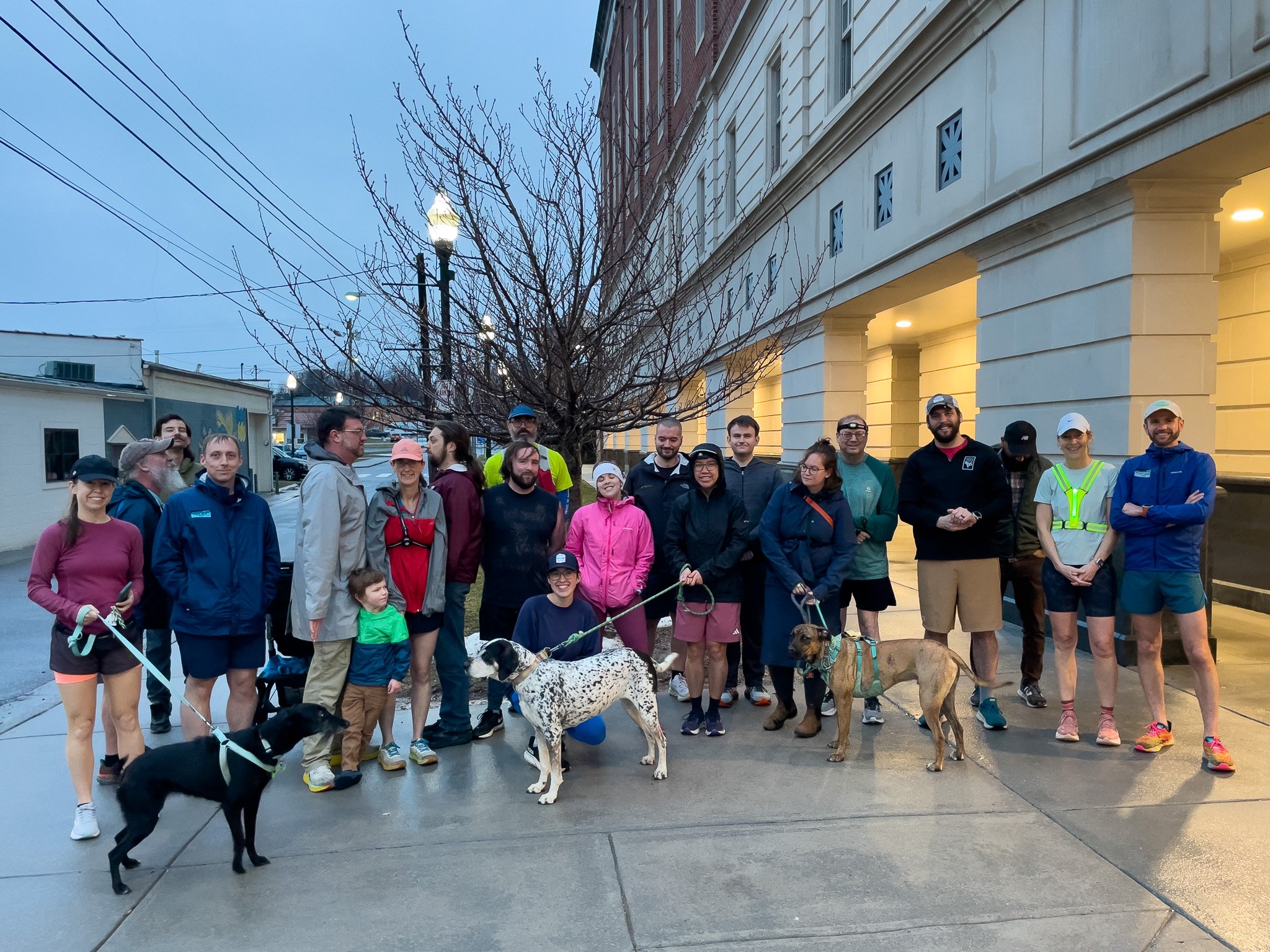 Group of people with dogs standing on sidewalk in front of a building during evening.