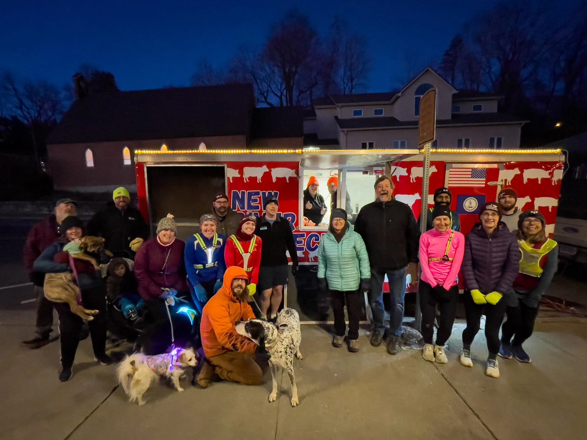 Group of people with dogs standing in front of a food truck decorated with cow images, American flag, and text at dusk.