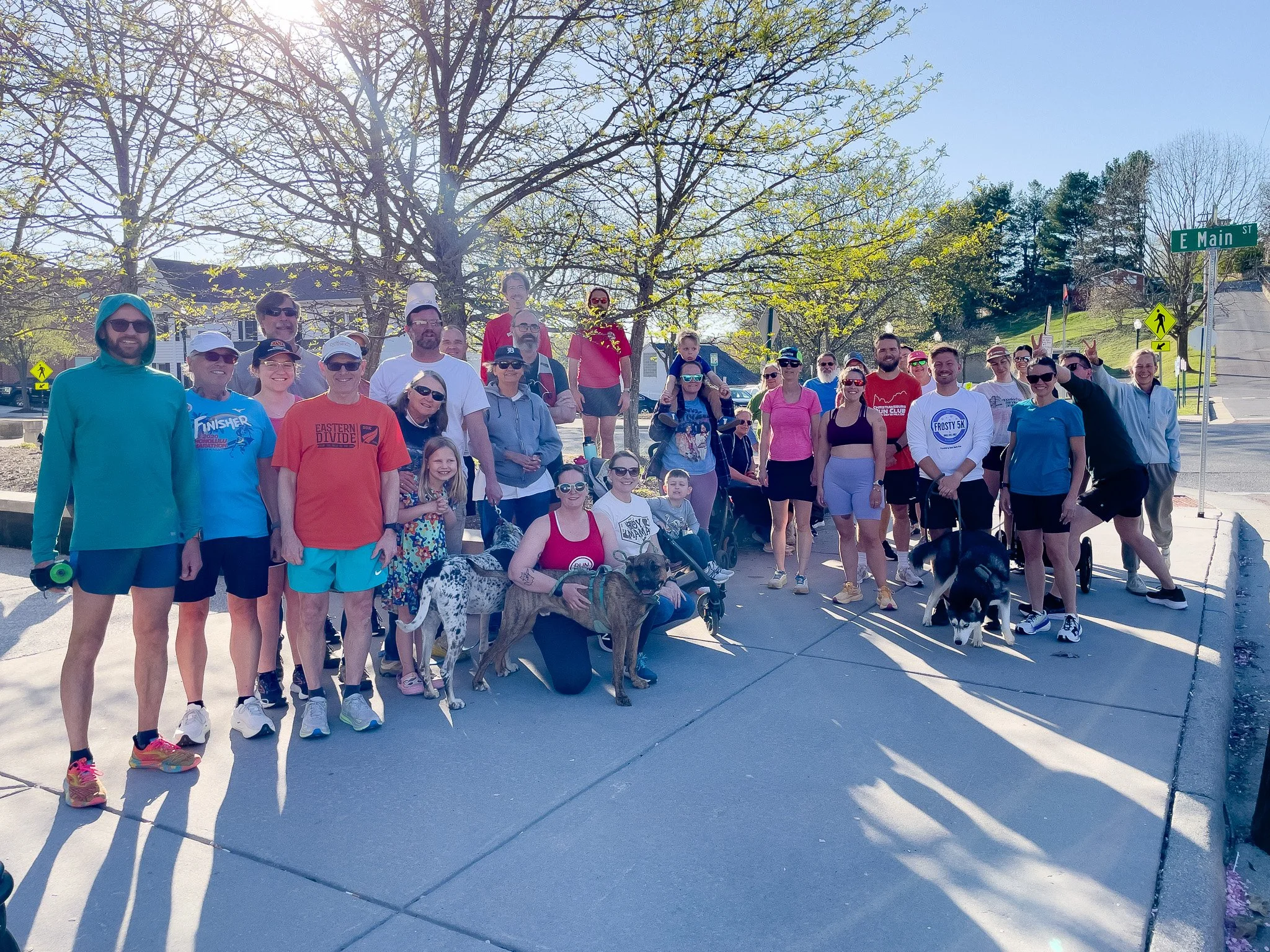 Group of people and dogs posing for a photo outdoors on a sunny day near a street intersection with signs including 'E Main St'. Some are wearing athletic clothing and sunglasses. The background features leafless and green trees and a hill.