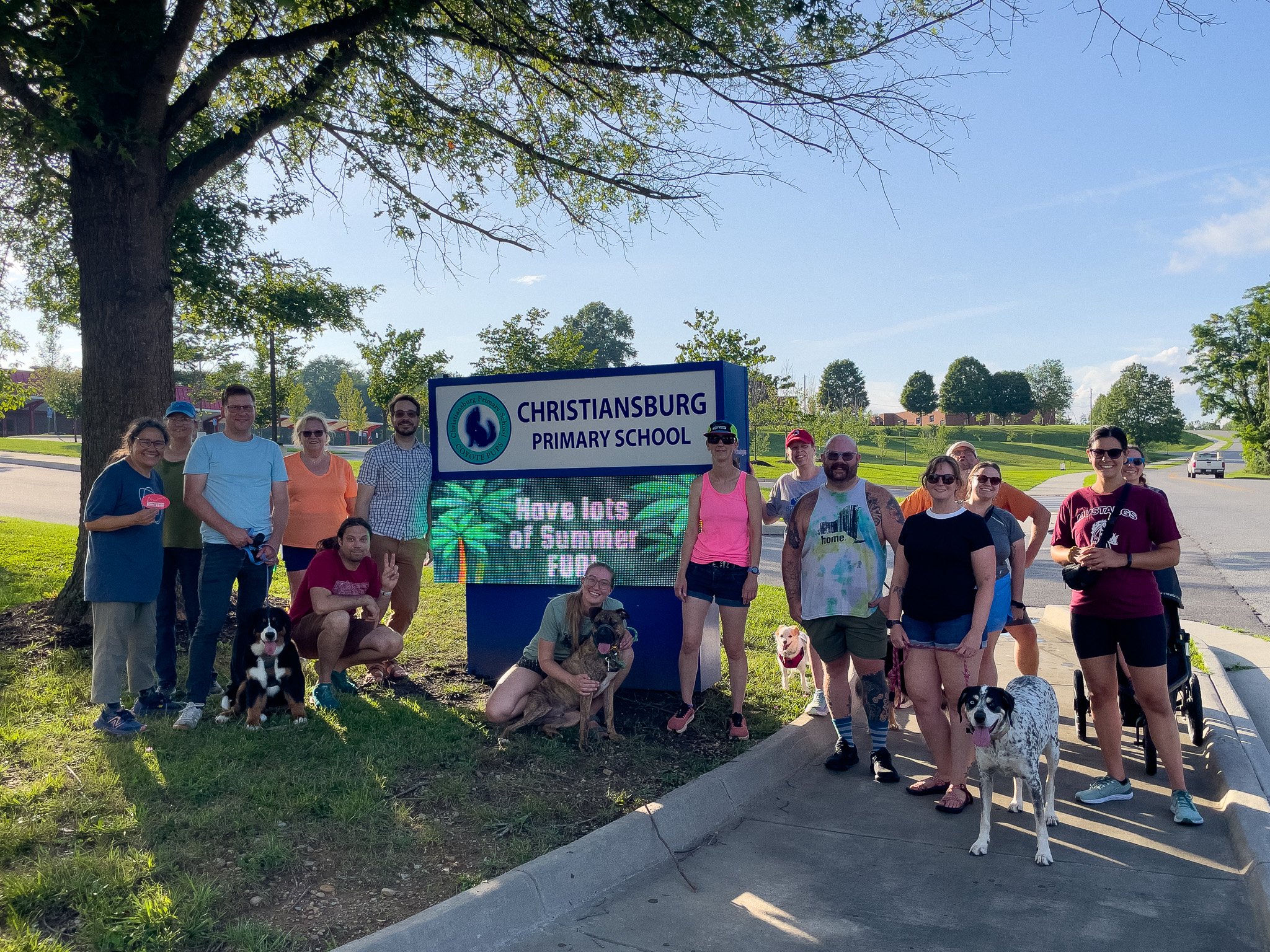 Group of people standing next to a sign for Christiansburg Primary School on a sunny day, with some dogs and trees in the background.