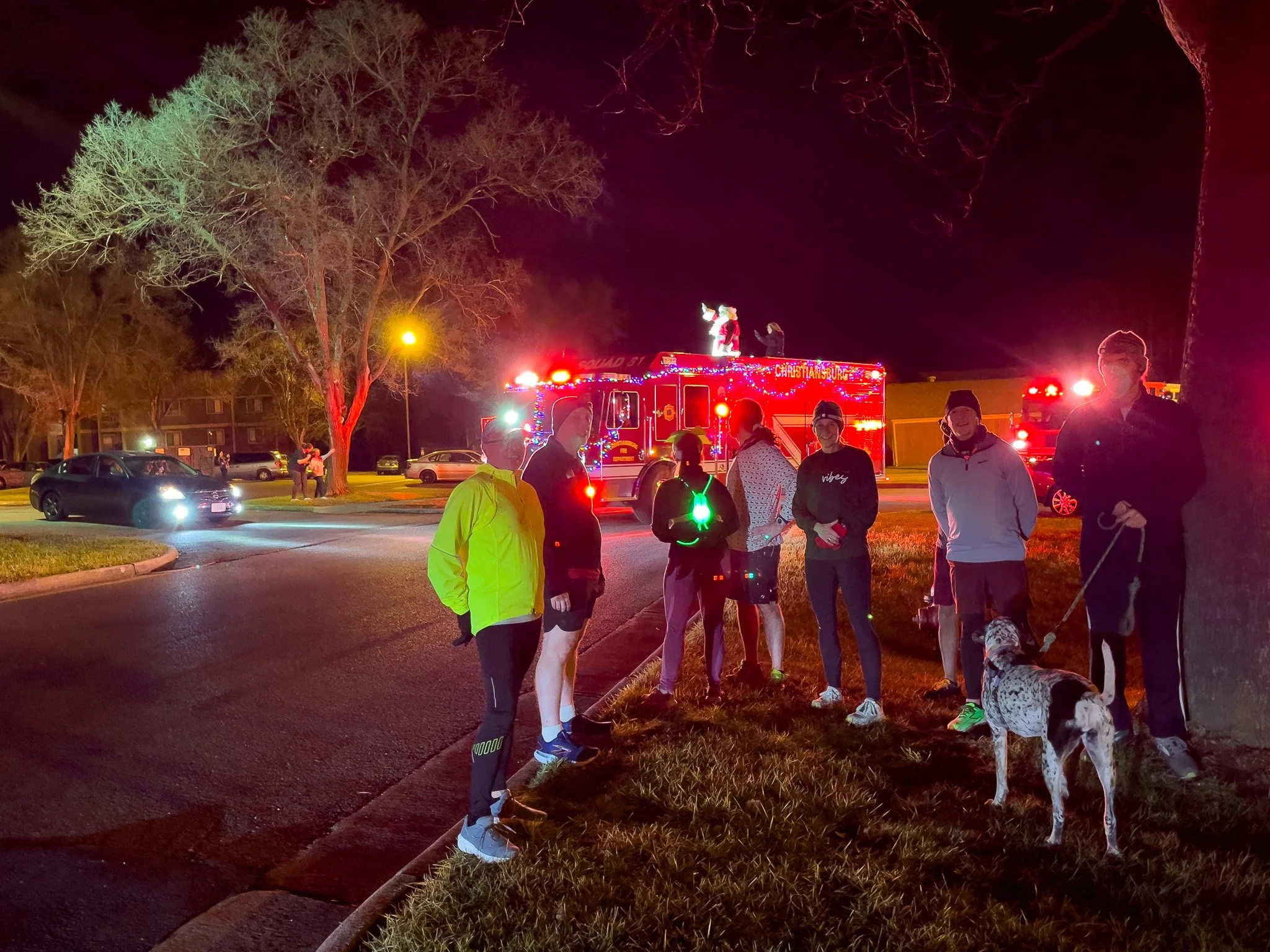Group of people standing on grass near street at night, with a fire truck in the background and Christmas lights on the fire truck.