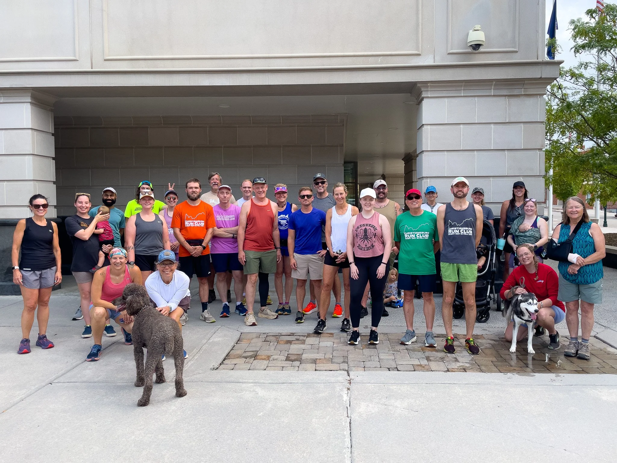 Group of people dressed in athletic clothing, standing outside a building, some with dogs. It appears to be a running club or outdoor fitness event.