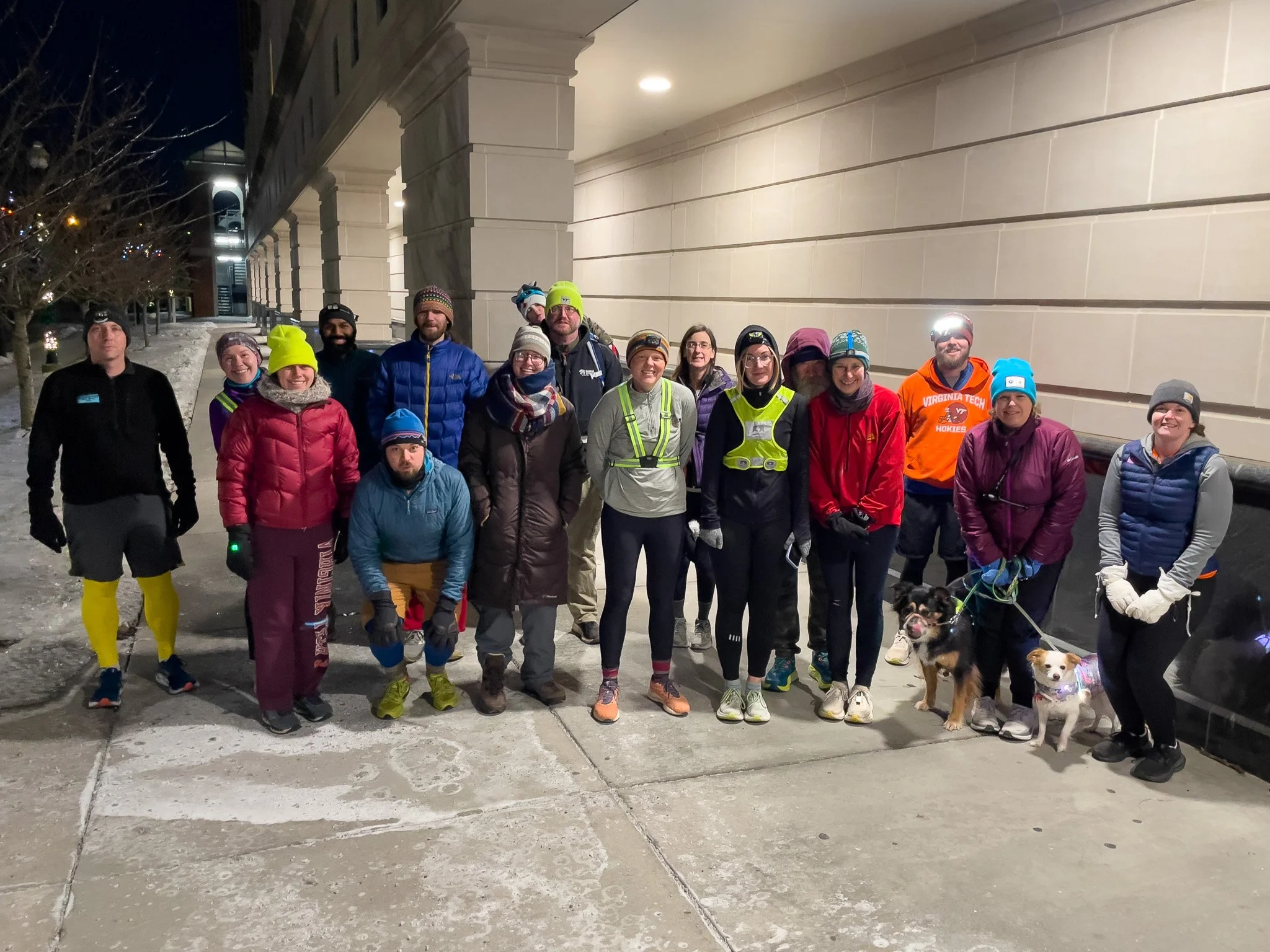 Group of runners dressed in athletic gear and winter clothing, standing under a building's overhang at night, some with headlamps on, accompanying two dogs on leashes.