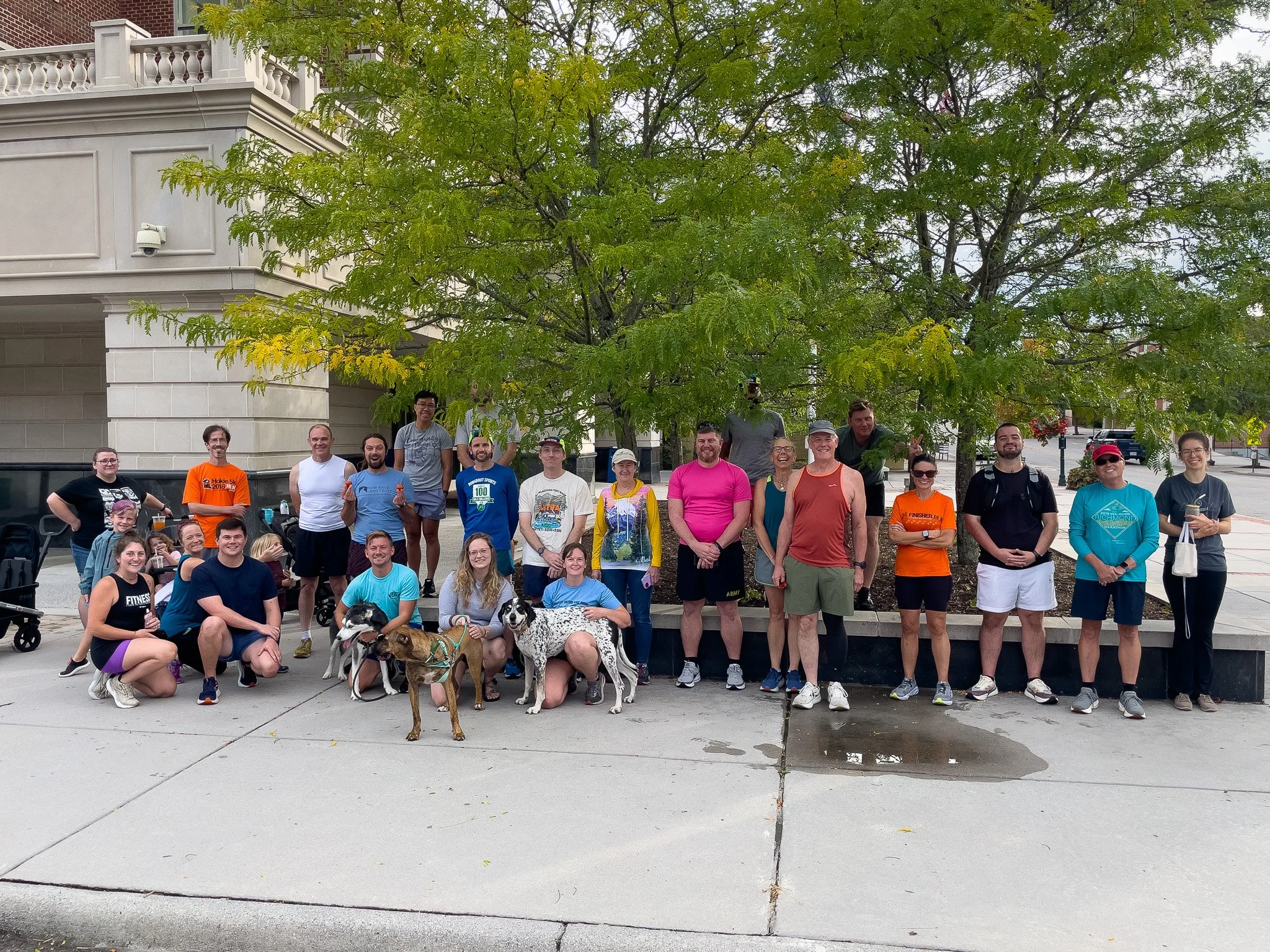 Group of people, some with dogs, gathered outdoors beneath a large green tree in front of a beige building, with some smiling and posing for the camera.