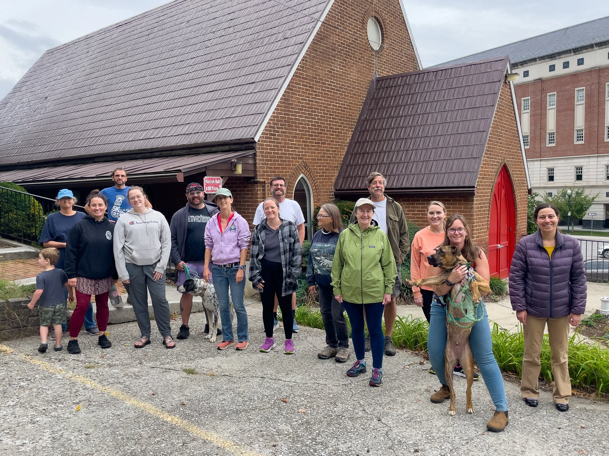 A group of people, including children and adults, standing outside a brick church with a red door. Some are smiling, and a woman is holding a large dog. The scene appears to be a community gathering or outing.