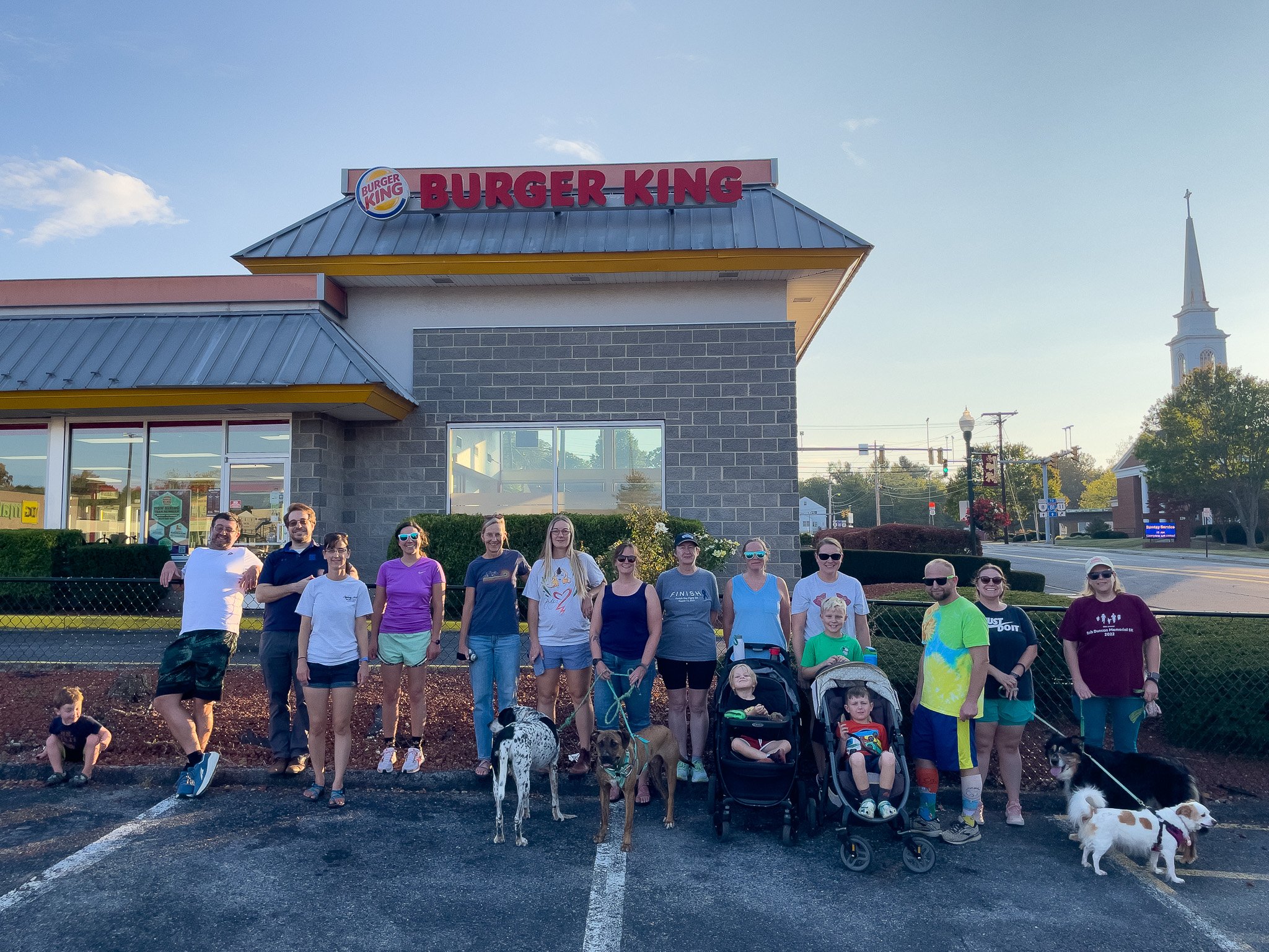 A group of people and dogs standing in front of a Burger King restaurant on a parking lot with a church steeple in the background.