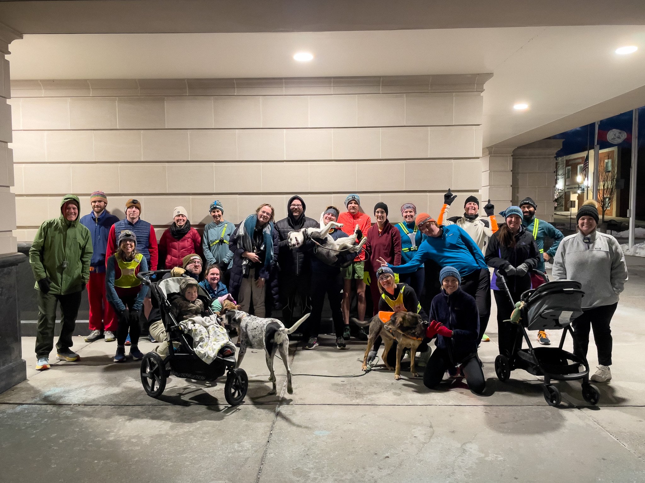 Group of people in winter clothing and running gear posing outdoors at night, with dogs and baby strollers.