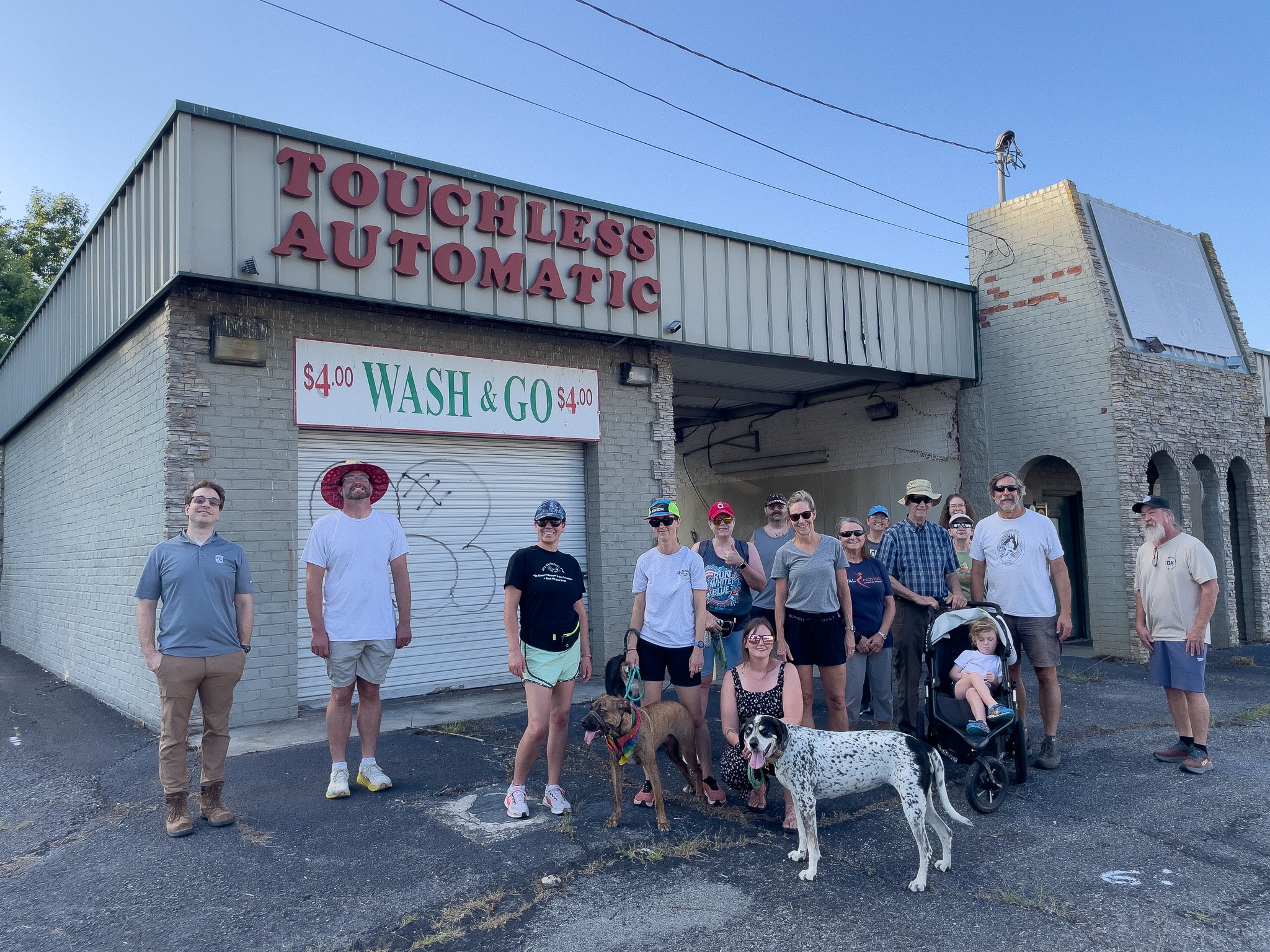 A group of people standing in front of a building with a sign that reads 'Touchless Automatic' and a smaller sign that says '$4.00 Wash & Go'. There are two dogs and a child in a stroller among the group.