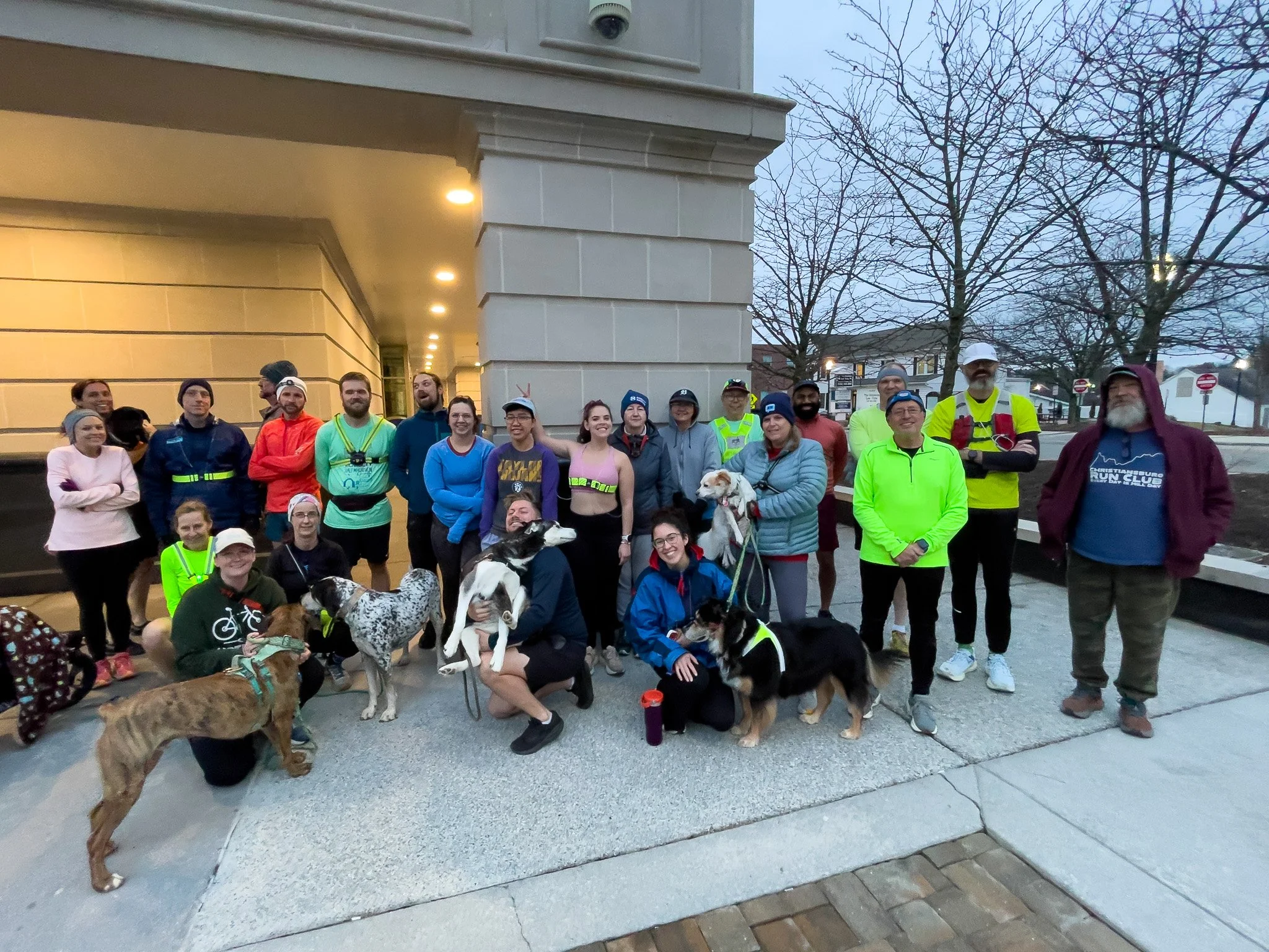 Group of people and dogs gathering outdoors at dusk, some in running attire, in front of a building with trees in the background.