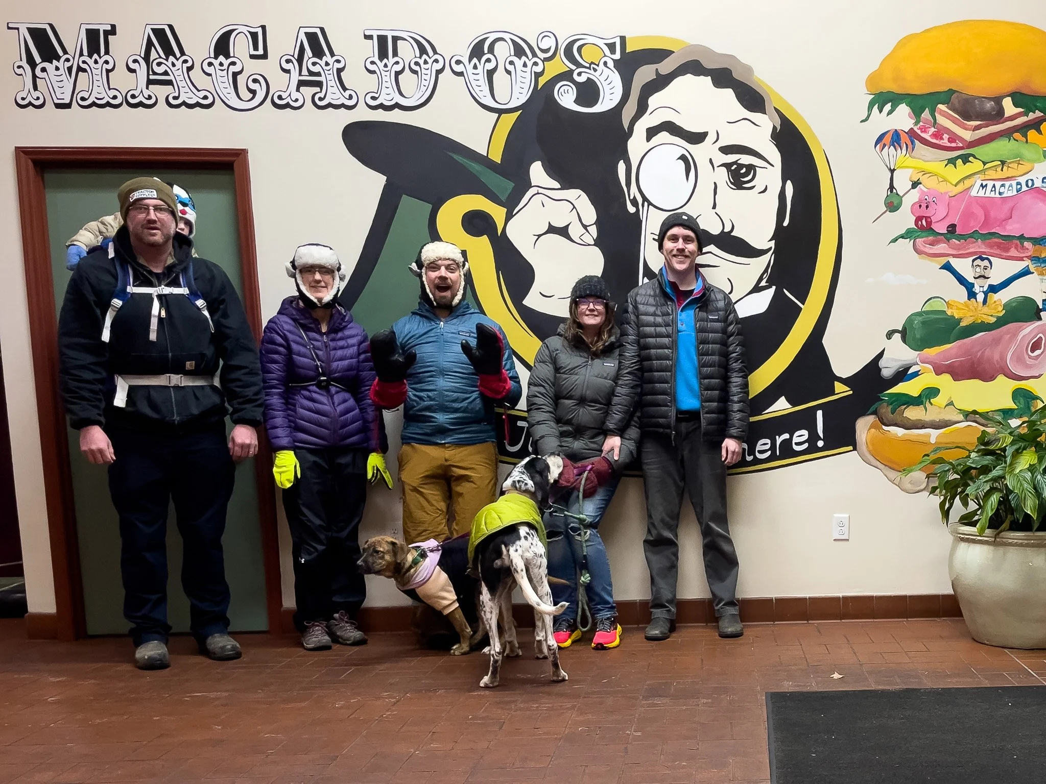 A group of five people and two dogs posing inside a restaurant with a mural of a man pointing a finger and a large burger on the wall behind them. The people are dressed in winter outdoor gear.
