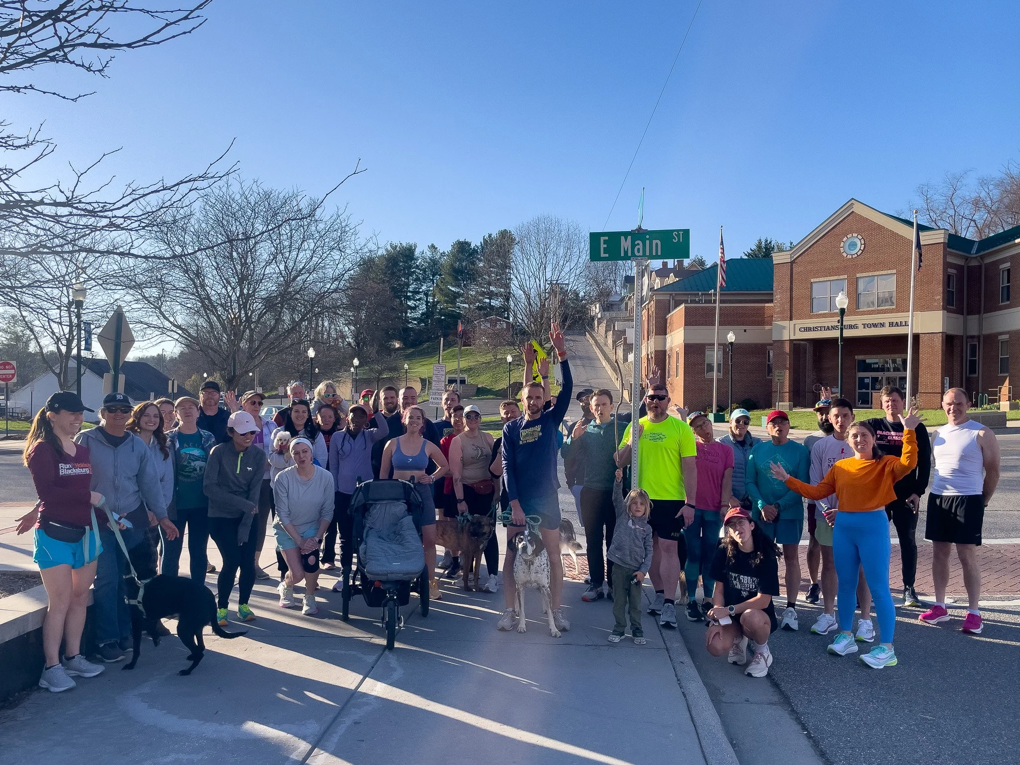A large group of people gathered outdoors at an intersection on a sunny day, some wearing athletic clothes, with a building labeled 'Christiansburg Town Hall' in the background and a street sign that reads 'E Main St.'