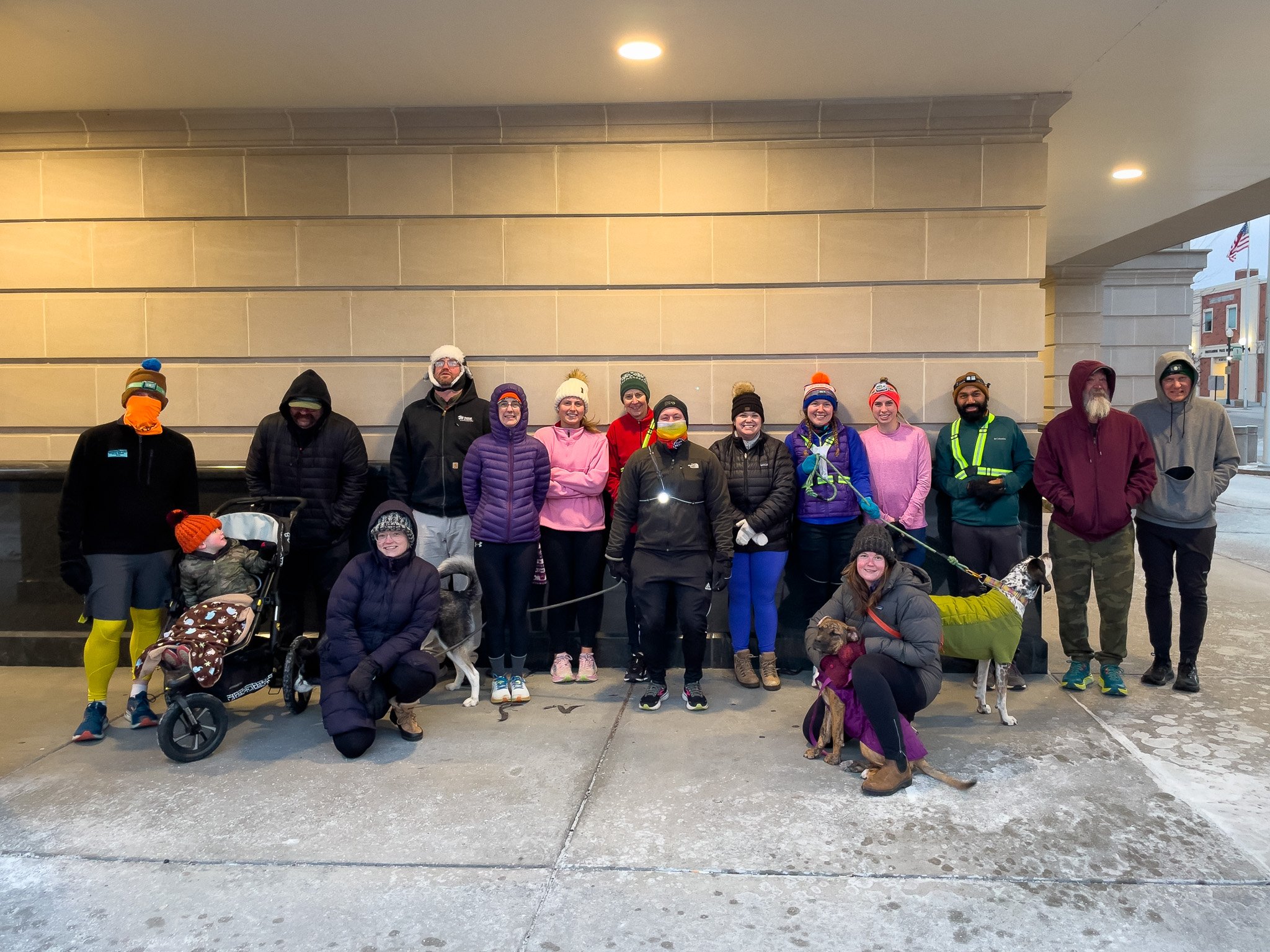 Group of people dressed in cold weather gear standing outside on a winter day, some with dogs and strollers, in front of a beige building with an American flag in the background.