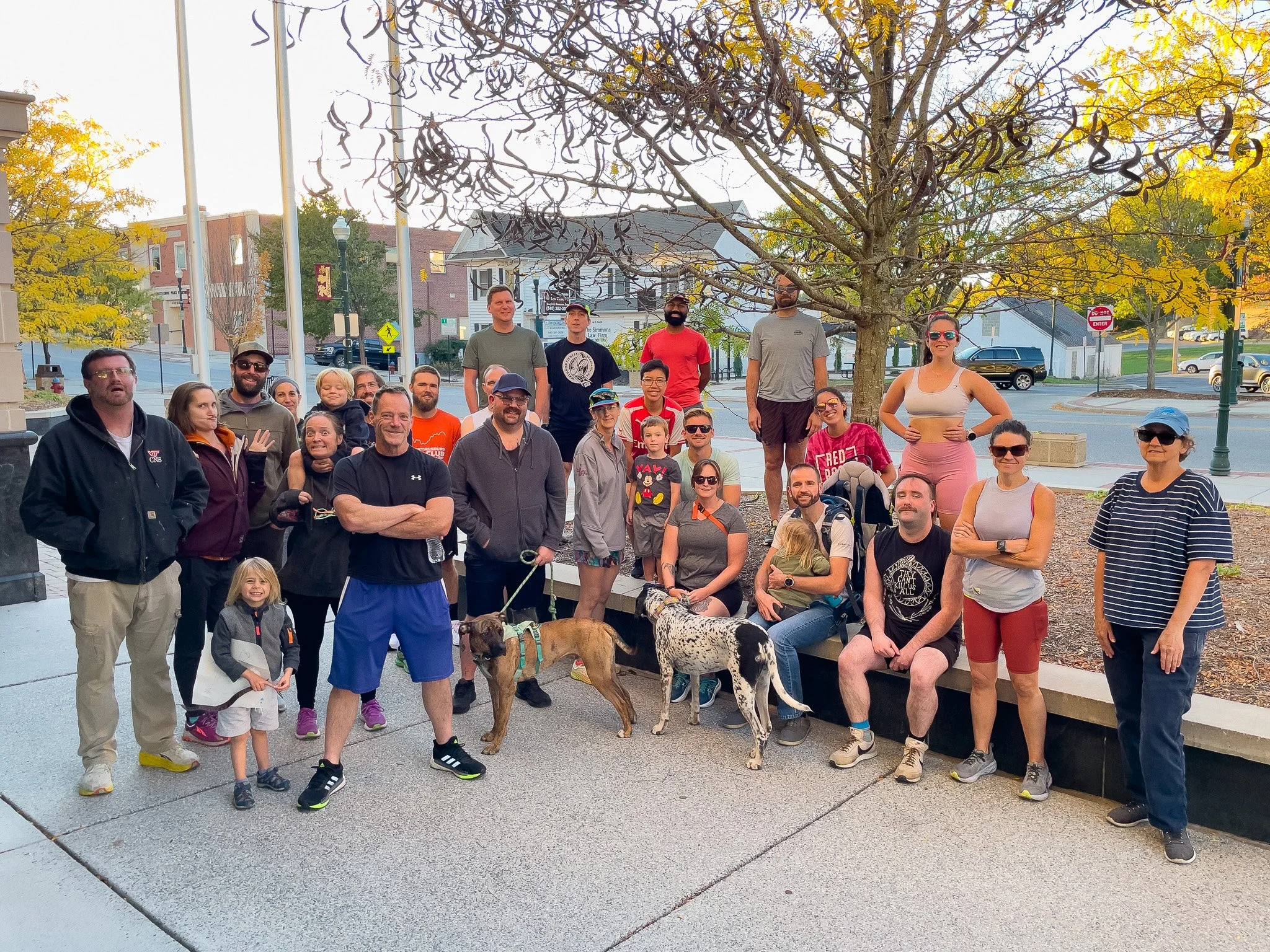 A group of people, including children and adults, posing outdoors on a sidewalk with trees and buildings in the background.