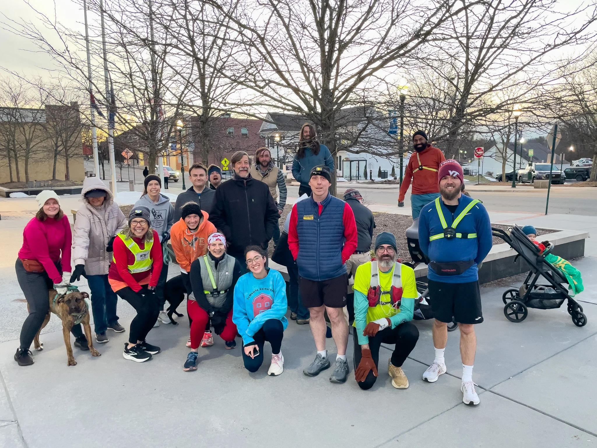 Group of people dressed in athletic gear standing outside on a cold day, some wearing reflective vests, with dogs and a stroller, in an urban park area with leafless trees and buildings in the background.