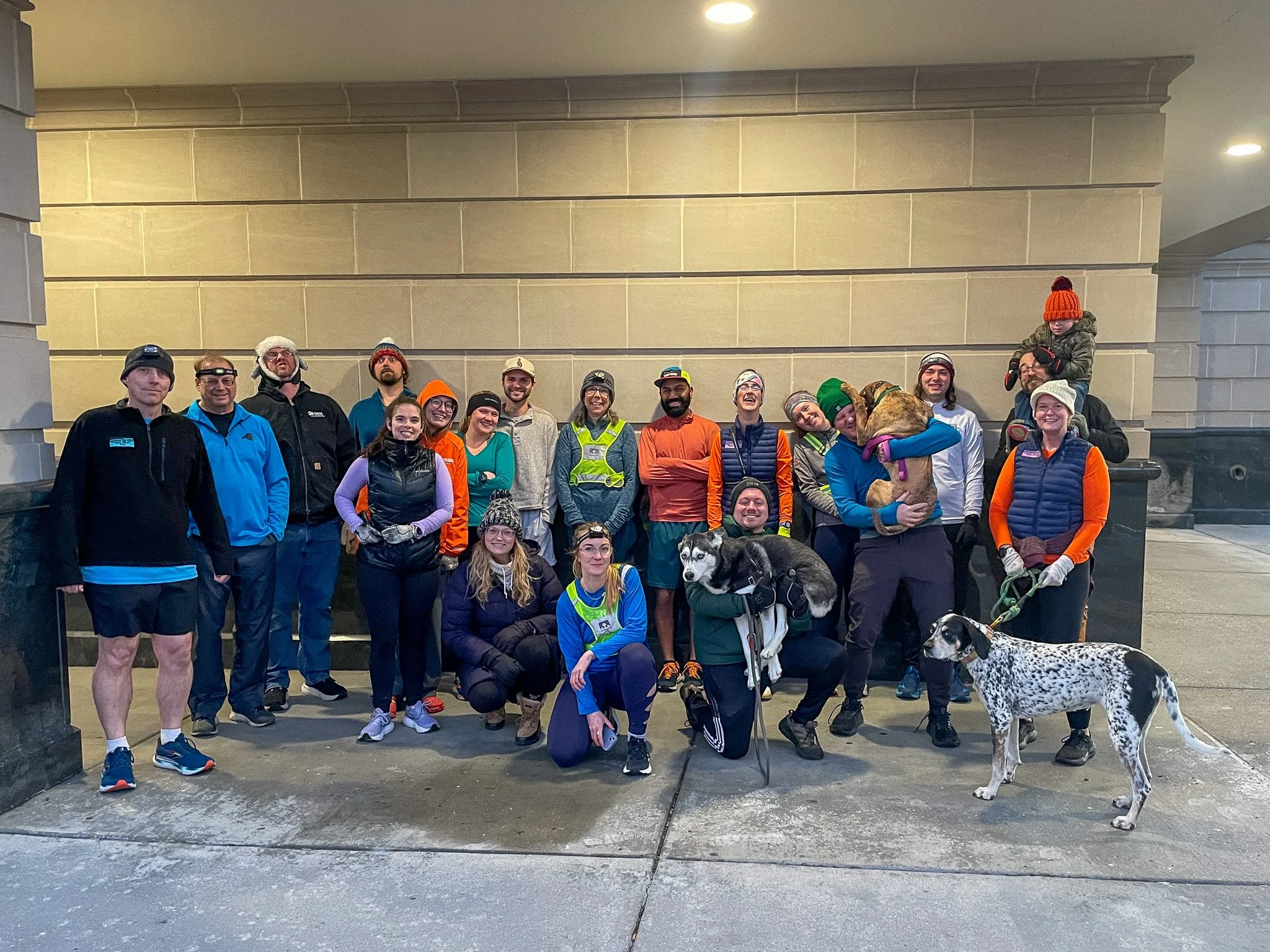 Group of people and dogs posing outside under a beige stone building wall at night, dressed in athletic and outdoor gear, some wearing hats and gloves.