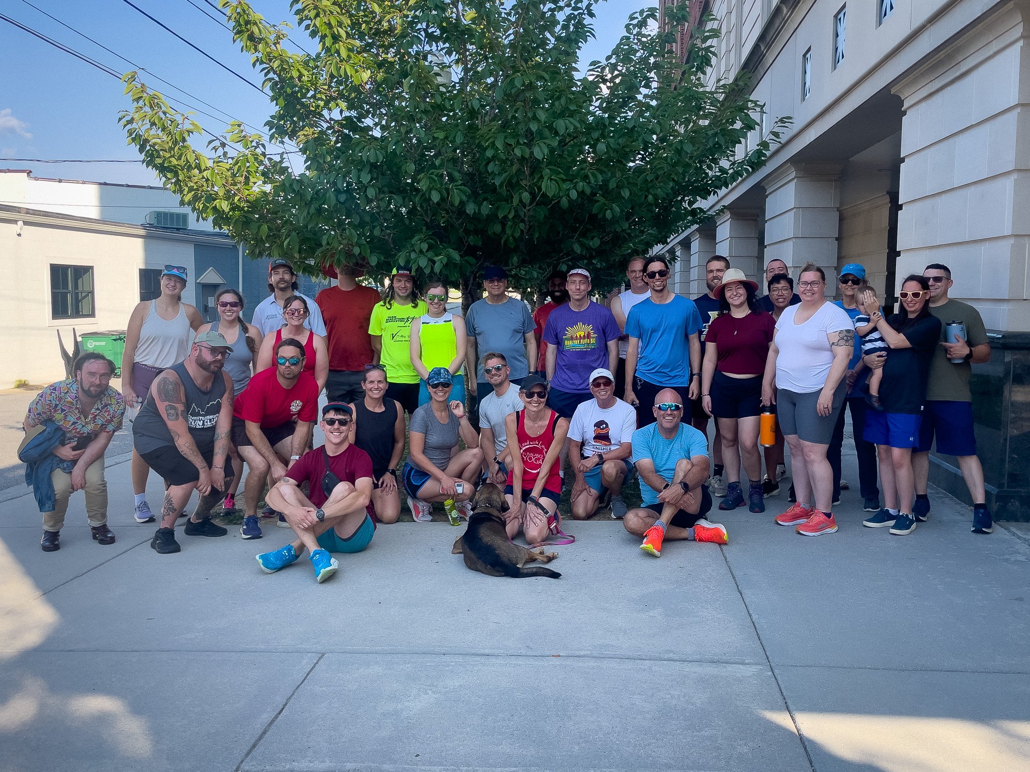 Group of people posing outdoors in front of a tree and building on a sunny day. Some are wearing athletic clothing and sunglasses. There is a dog lying on the ground in front of the group.