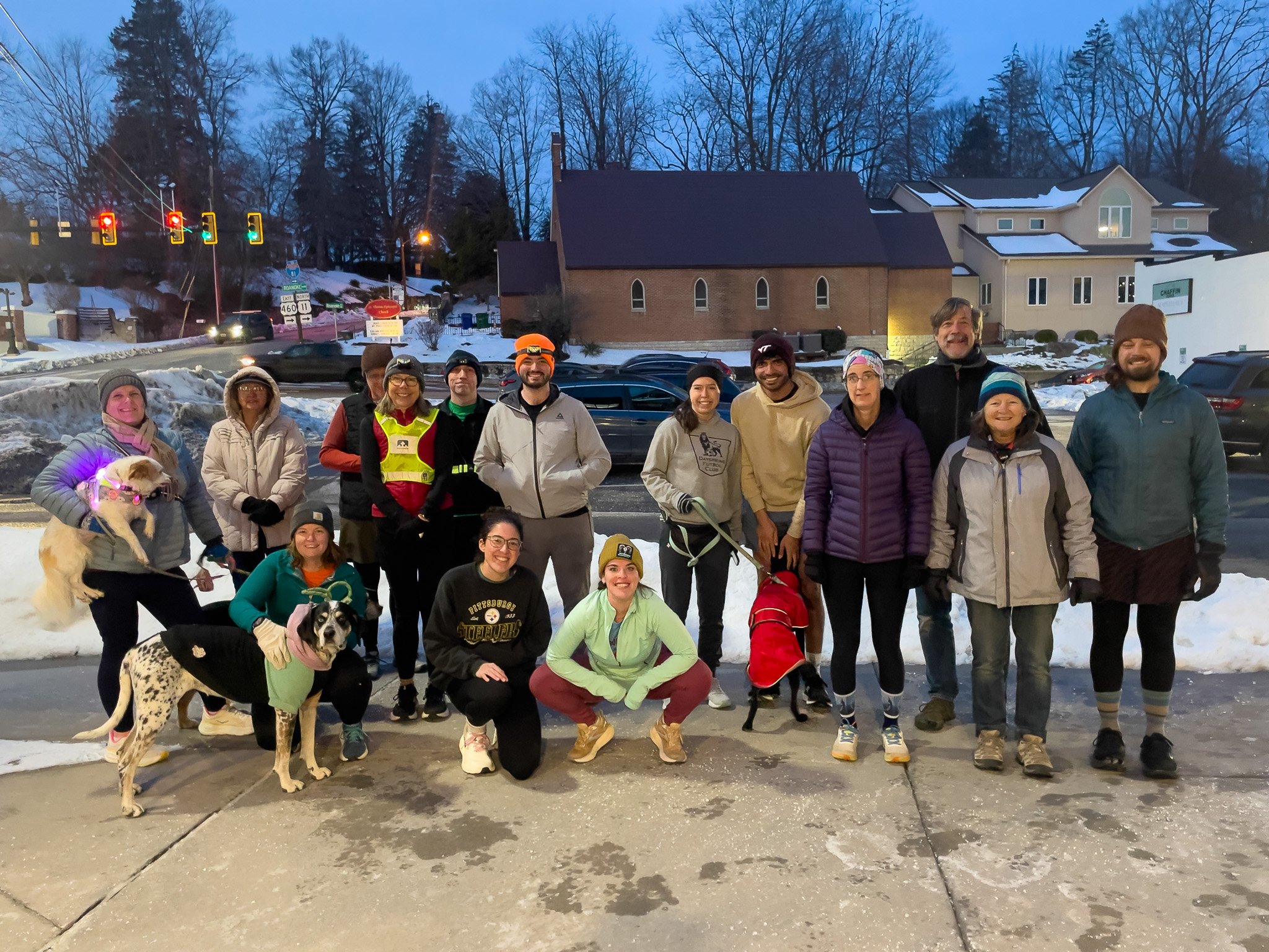 Group of 15 people and 2 dogs standing outdoors on a snowy sidewalk in front of a residential area with houses, trees, and a traffic light in the background during the evening.