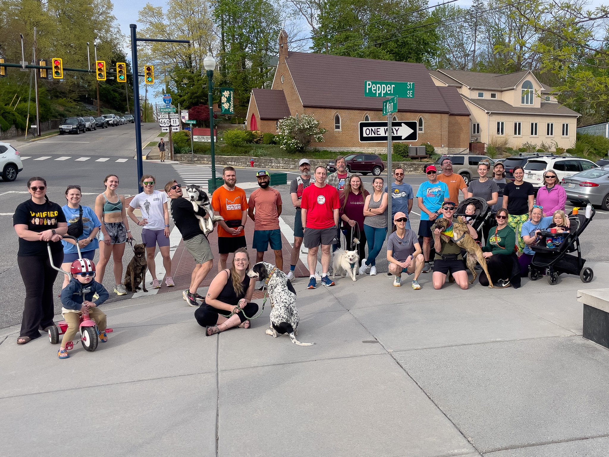 Group of people with dogs and children gathered on sidewalk at an intersection in a suburban neighborhood.