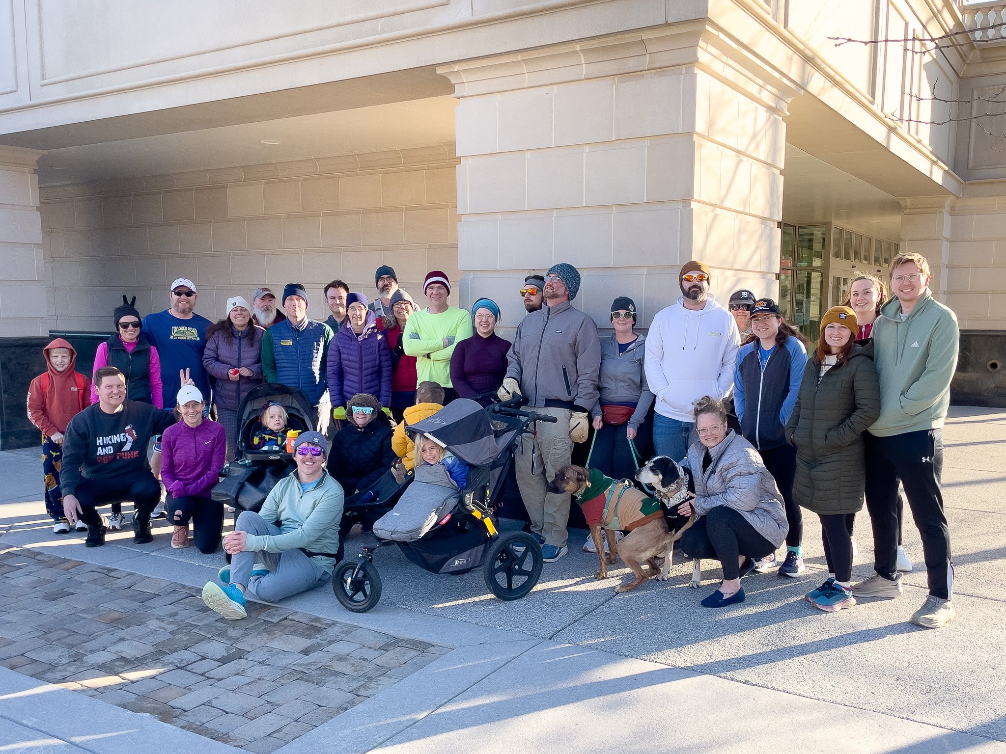 Group of people outdoors posing for a photo in front of a building, some with pets and strollers, dressed in casual and outdoor clothing.